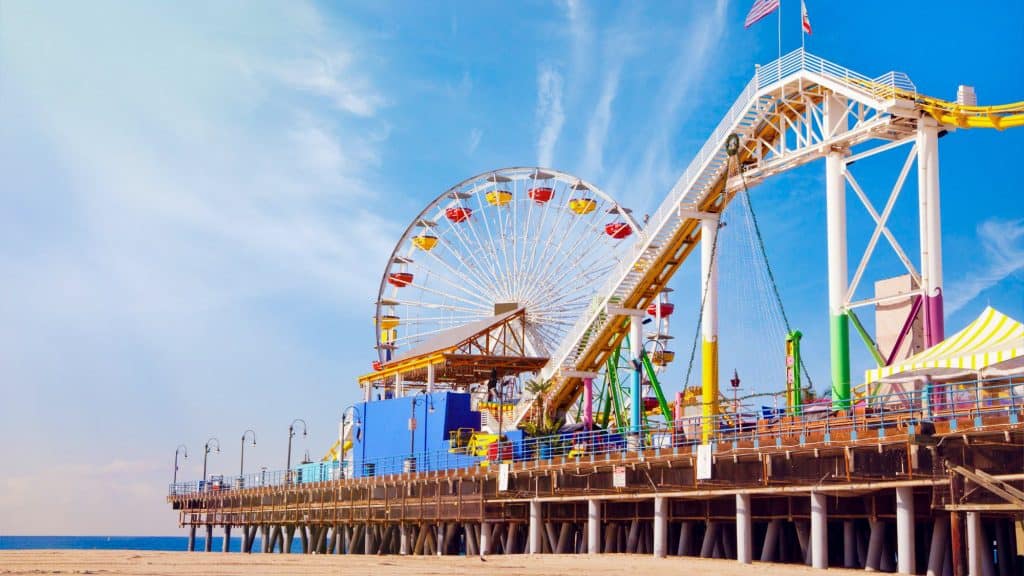 The vibrant Pacific Park amusement area on the Santa Monica Pier features a colorful Ferris wheel and roller coaster rising over the sandy beach under a bright blue sky.