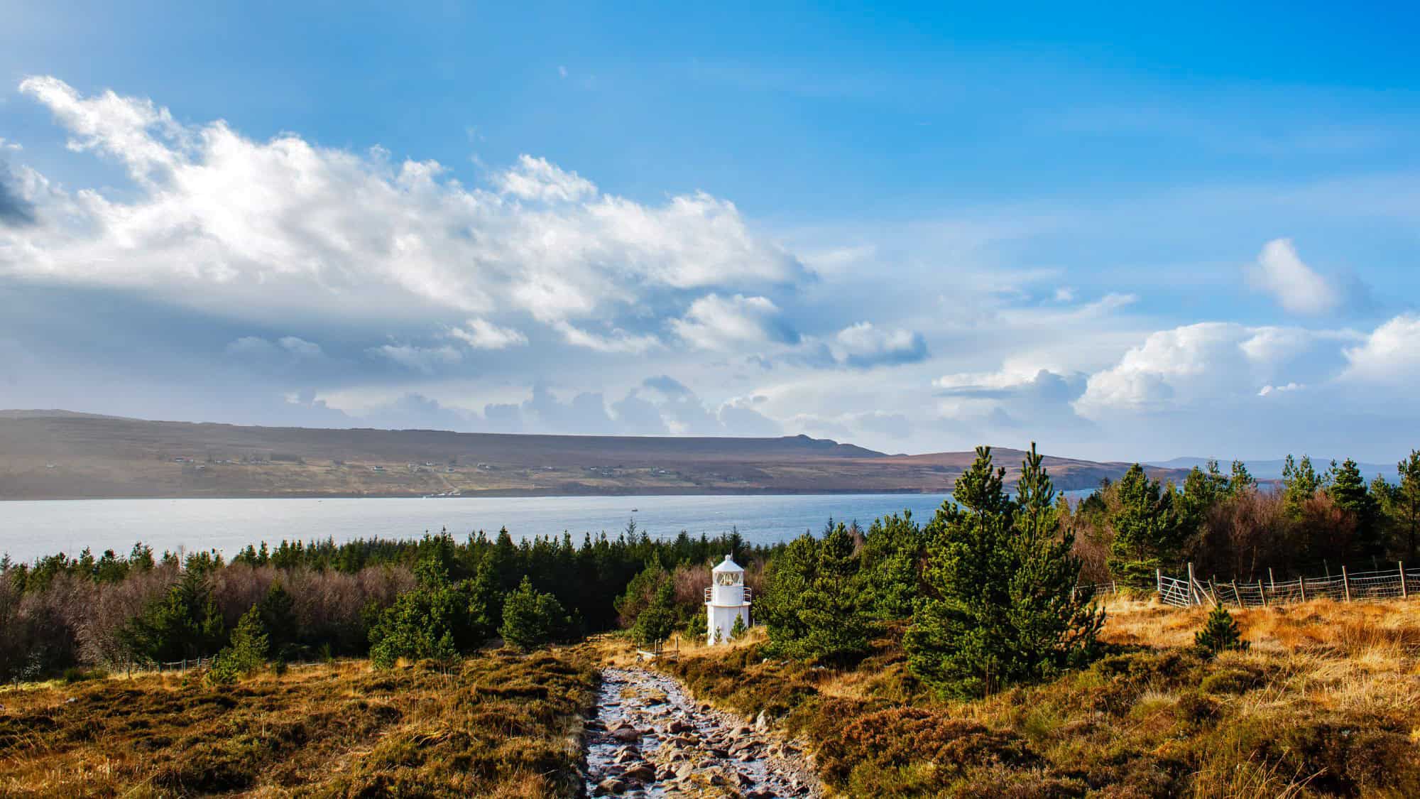 A tiny white lighthouse peeks out from a forest clearing near the coast in the Scottish Highlands, under a dramatic sky with rolling clouds and distant hills.