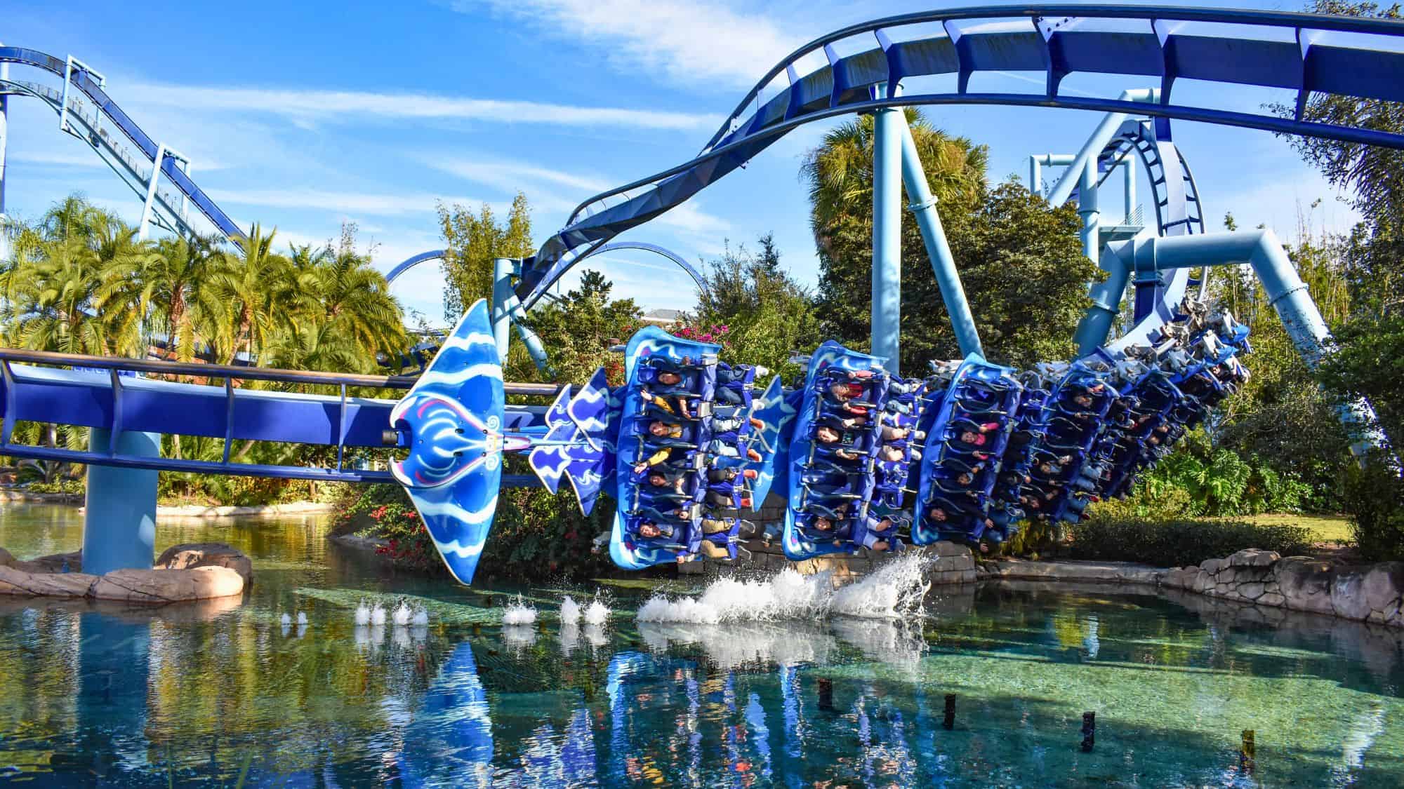 A manta ray-themed roller coaster soars over a reflective pool with riders suspended below the blue track and trees in the background.