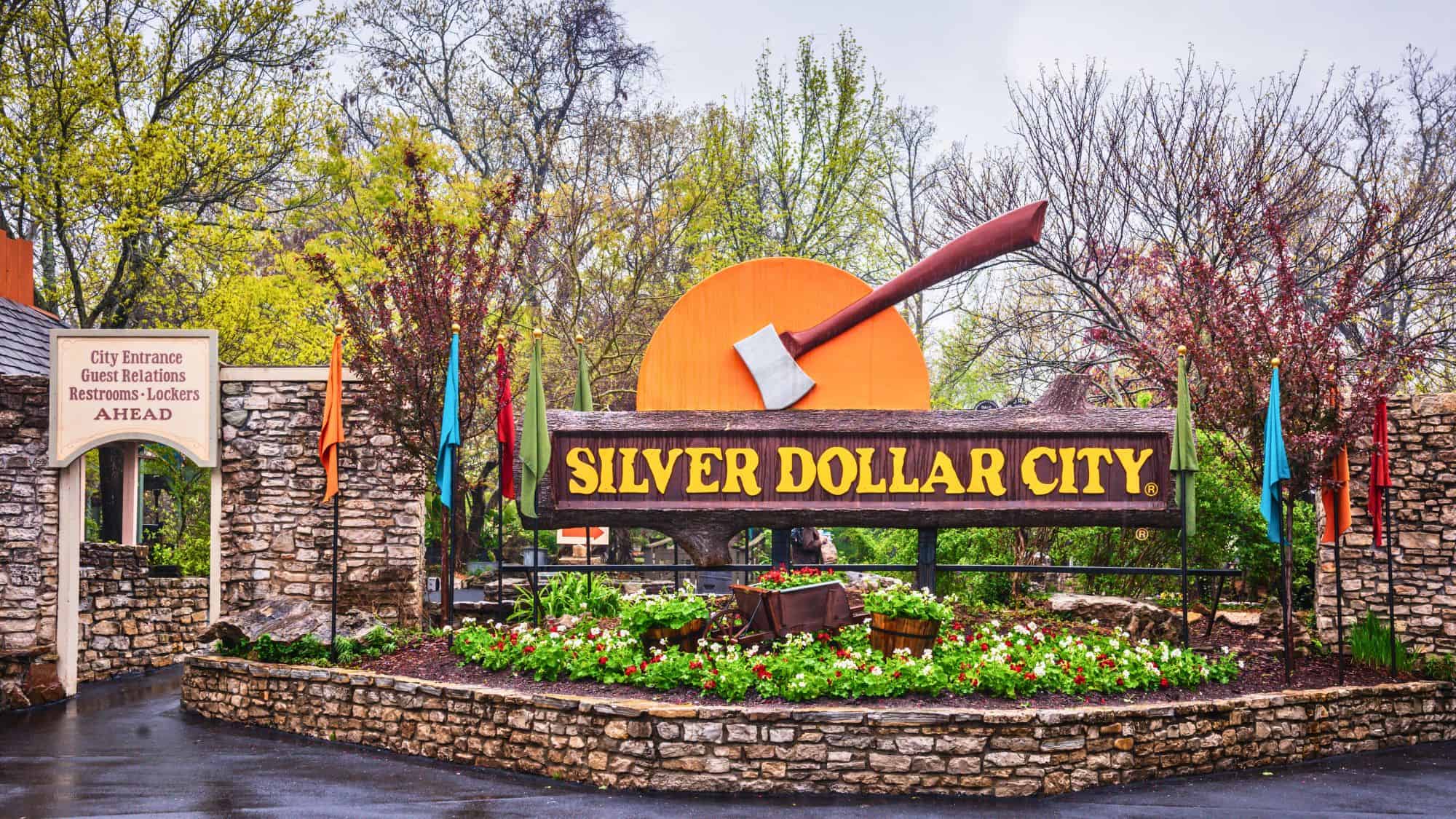 A rustic sign for Silver Dollar City features a giant axe embedded in a wooden log with colorful flags and blooming flowers in front.