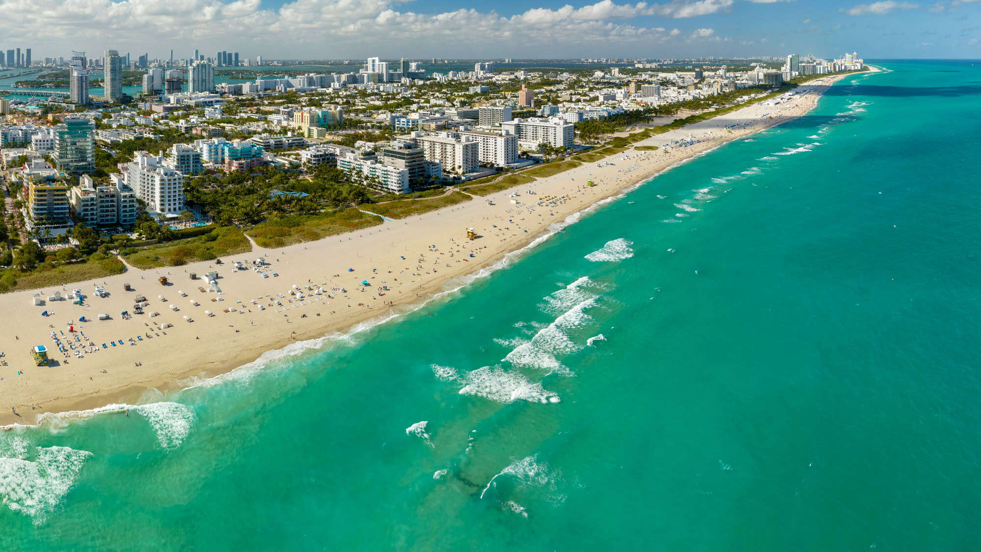 An expansive aerial of Miami’s South Beach shows rows of high-rise buildings and umbrellas lining the wide sandy shore beside turquoise waves.
