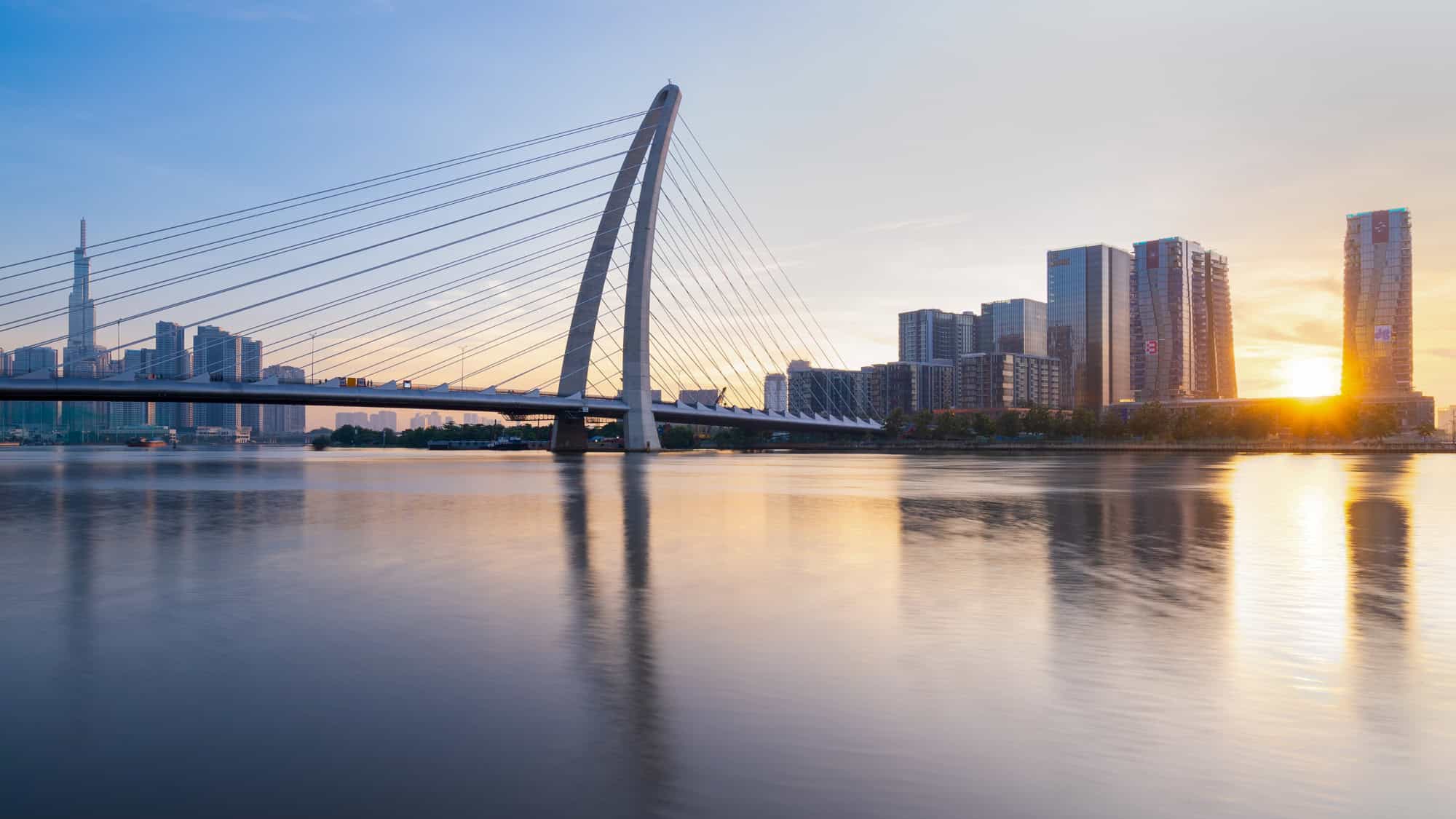 A sleek, modern cable-stayed bridge stretches across the Saigon River at sunrise, reflecting the glowing skyline and soft golden light of a new day.