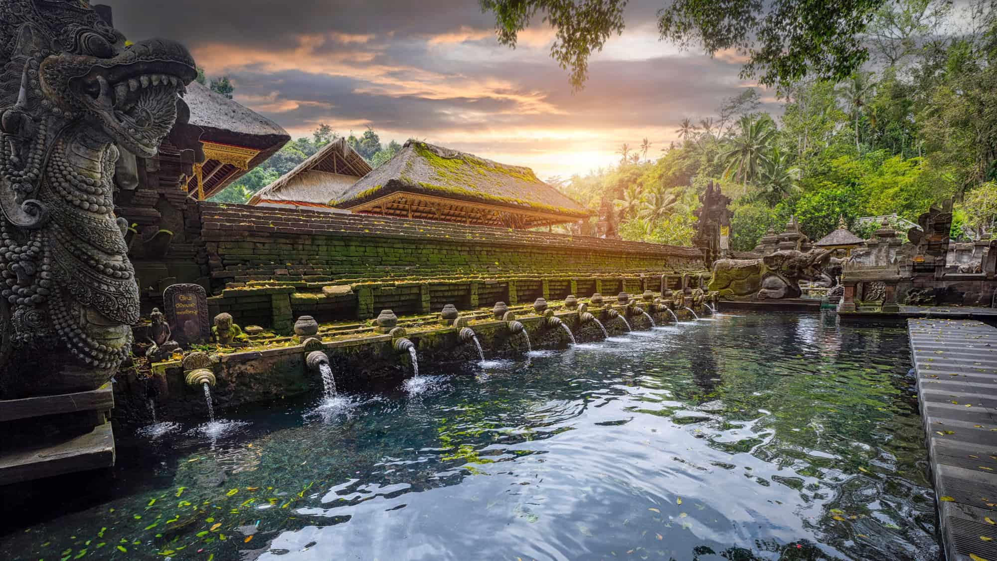 Water flows from intricately carved stone spouts into a tranquil pool at a Balinese temple complex, surrounded by moss-covered architecture and lush jungle, with warm sunset light in the background.