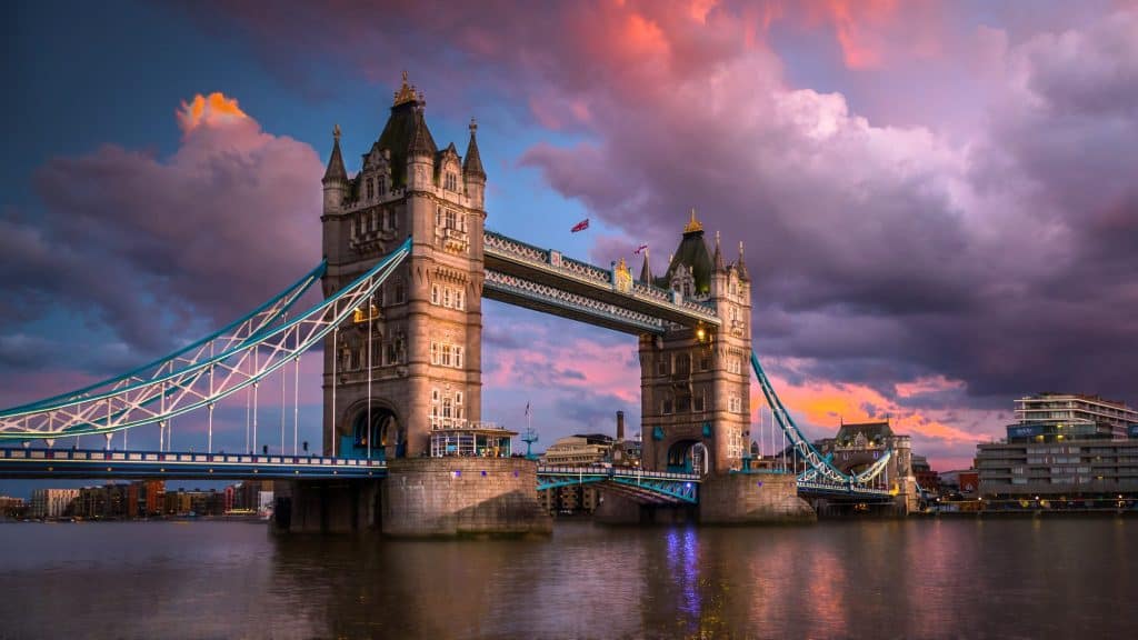 London’s Tower Bridge stands tall at sunset, lit warmly beneath a dramatic sky filled with purples and pinks, with reflections shimmering on the Thames River.