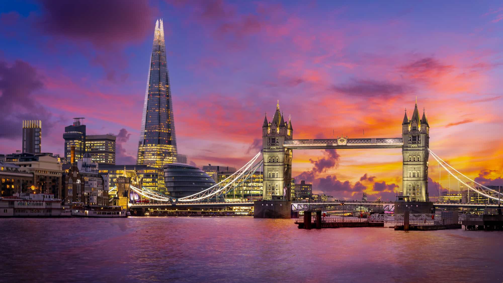 London’s Tower Bridge lights up at dusk against a backdrop of the illuminated Shard and modern skyline, under a vivid purple and orange sunset.