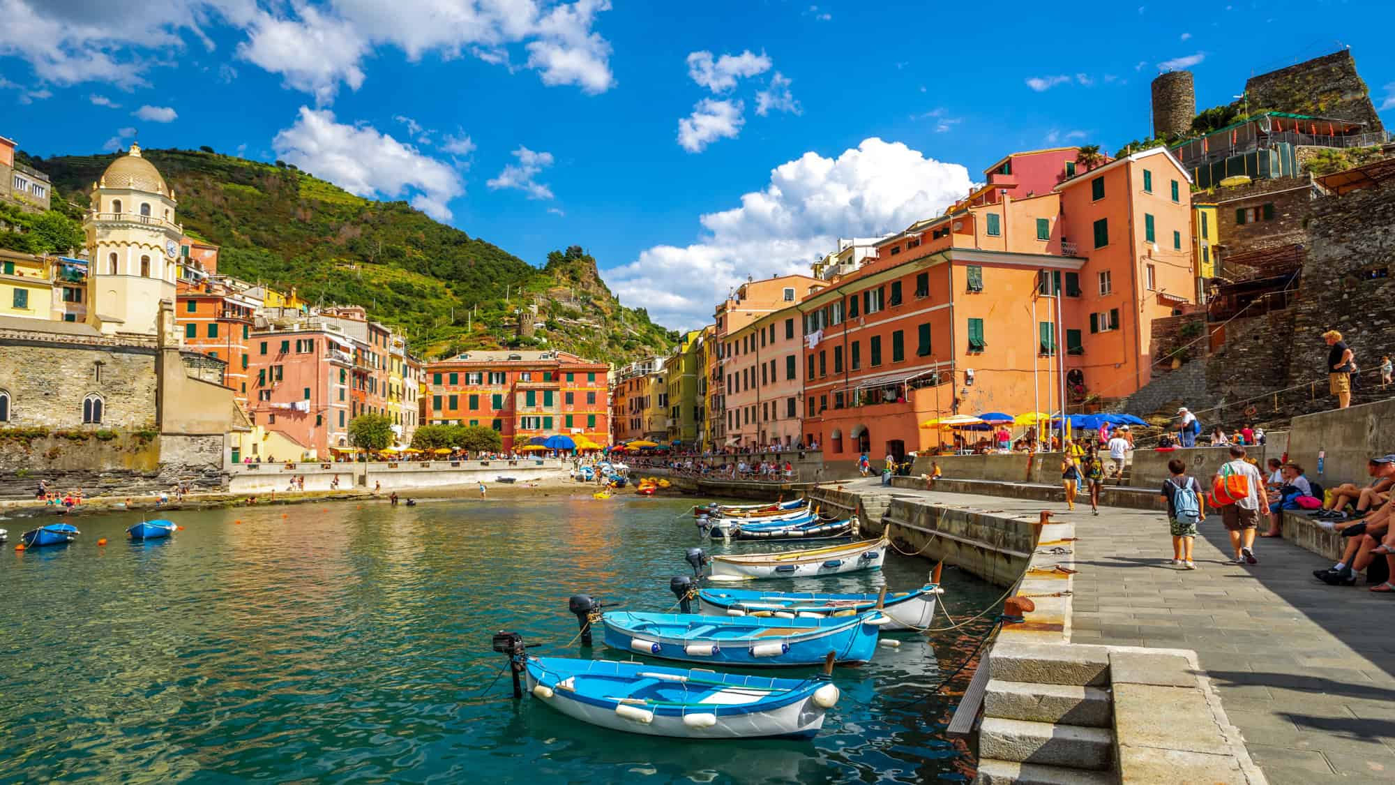 Vibrant pastel buildings line the harbor of Vernazza in Italy’s Cinque Terre, with small boats docked along the edge and tourists strolling the sunny promenade.