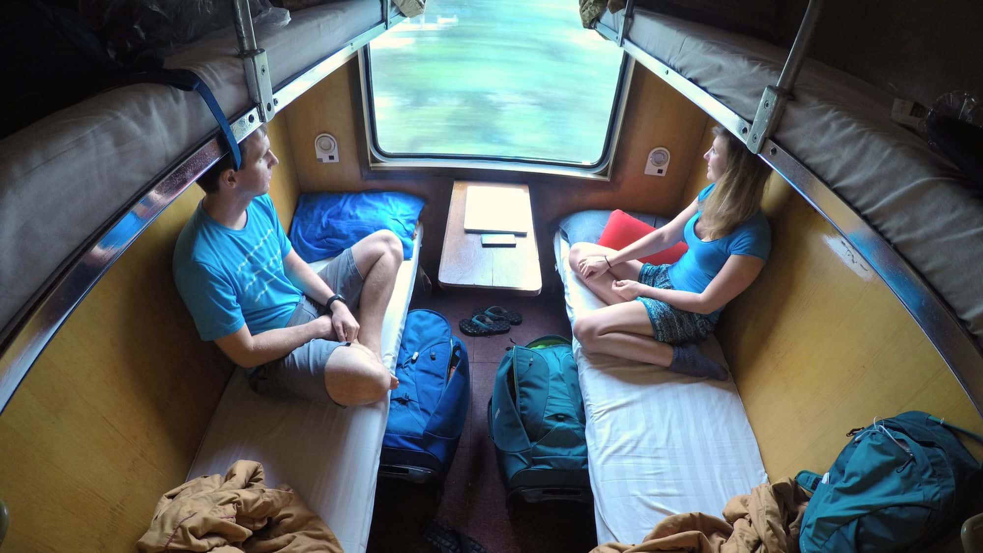 A cozy train compartment with two people seated on opposite bunks, surrounded by travel gear. The central window shows blurred greenery, suggesting motion and a relaxed travel experience.
