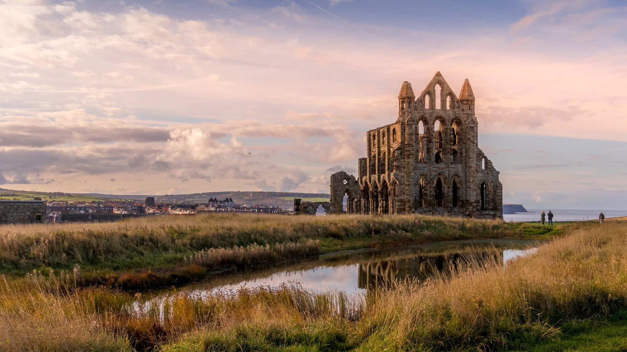 Ruins of Whitby Abbey sit on a grassy cliffside overlooking the North Sea at dusk, with a reflective pond in the foreground and soft pastel clouds stretching across the sky.