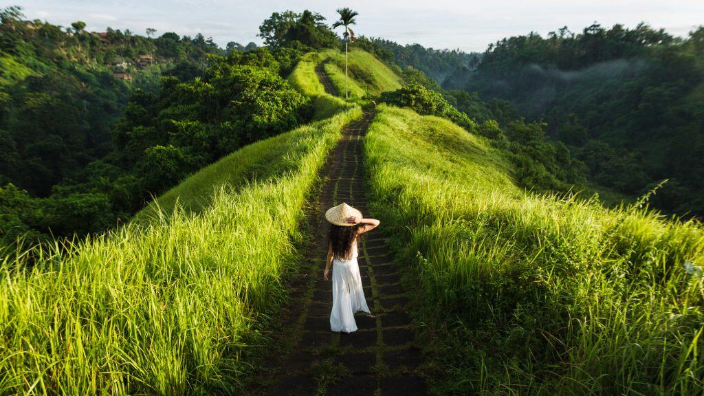 A woman in a white dress and straw hat walks along the narrow ridge of Campuhan Ridge Walk, flanked by tall green grass and tropical forested valleys.