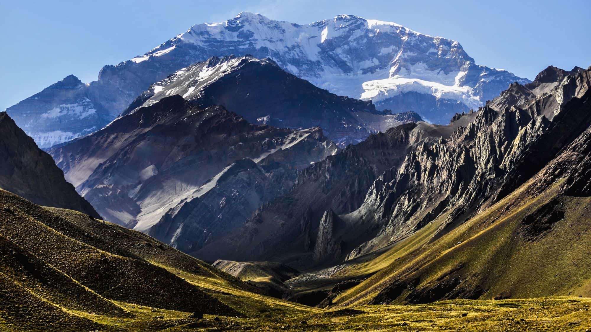 A dramatic view of jagged mountain ridges leading up to a towering snow-capped summit under a clear sky.
