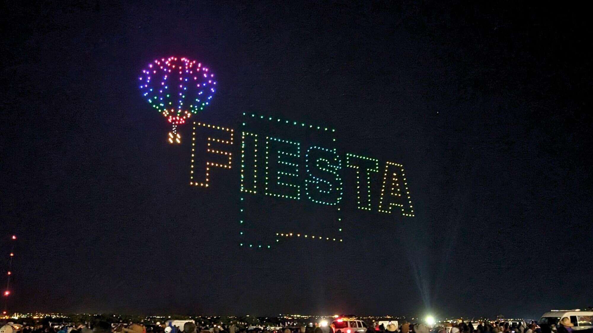 A nighttime drone display forms a colorful hot air balloon and the word “Fiesta” glowing above a large crowd.