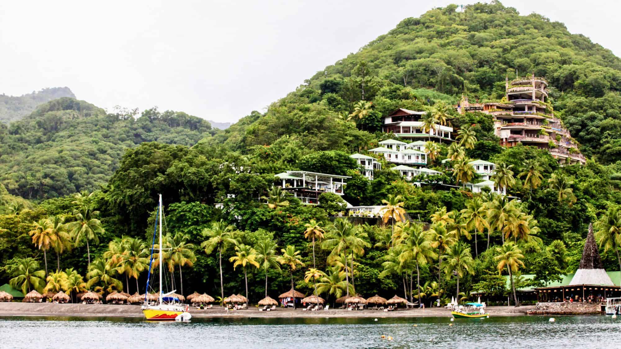 A lush green mountainside is dotted with tropical villas and resorts overlooking a beach lined with palm trees and thatched umbrellas.