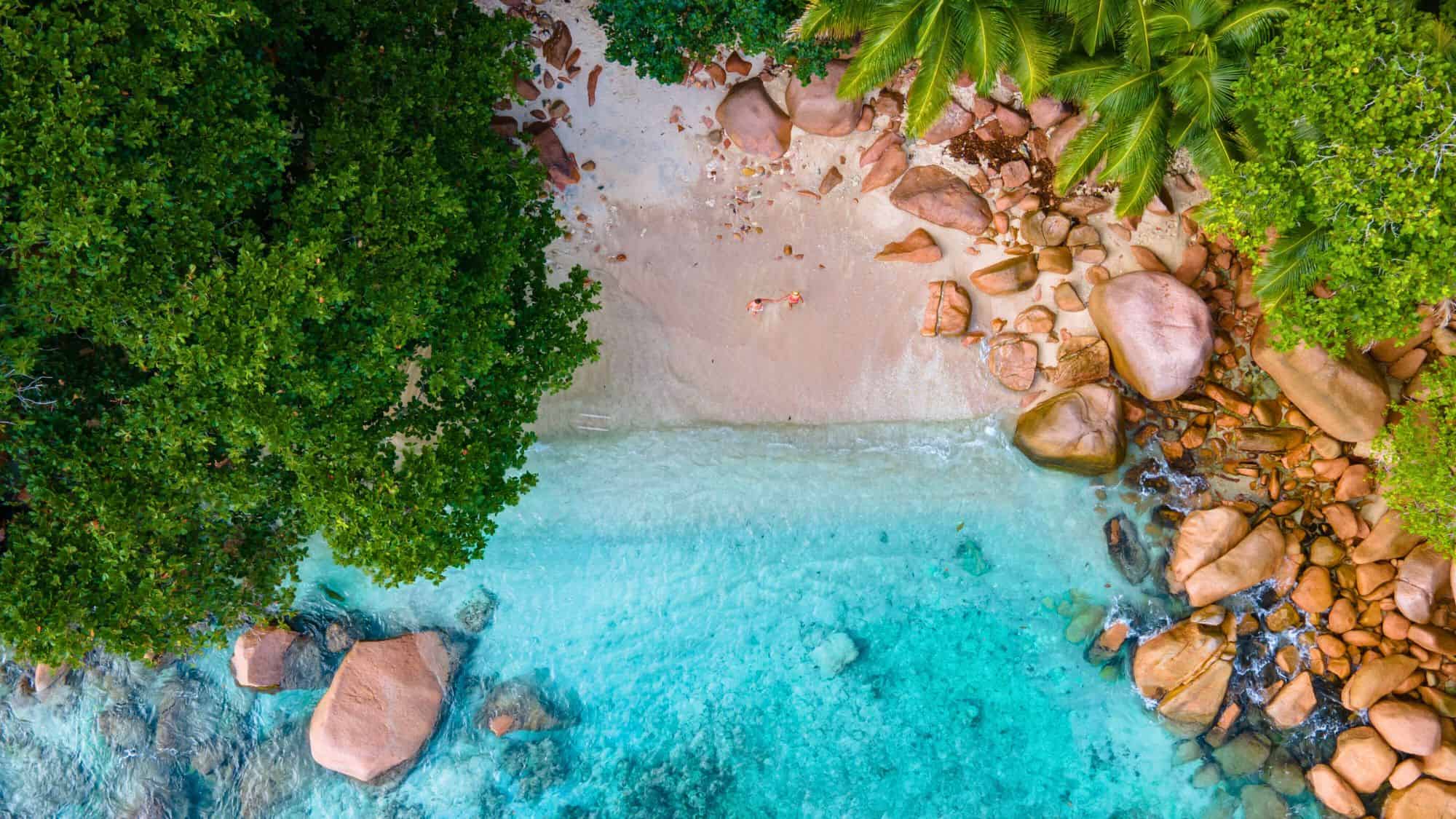 Overhead view of a secluded cove with turquoise water, large granite boulders, and two people relaxing on the soft sand.
