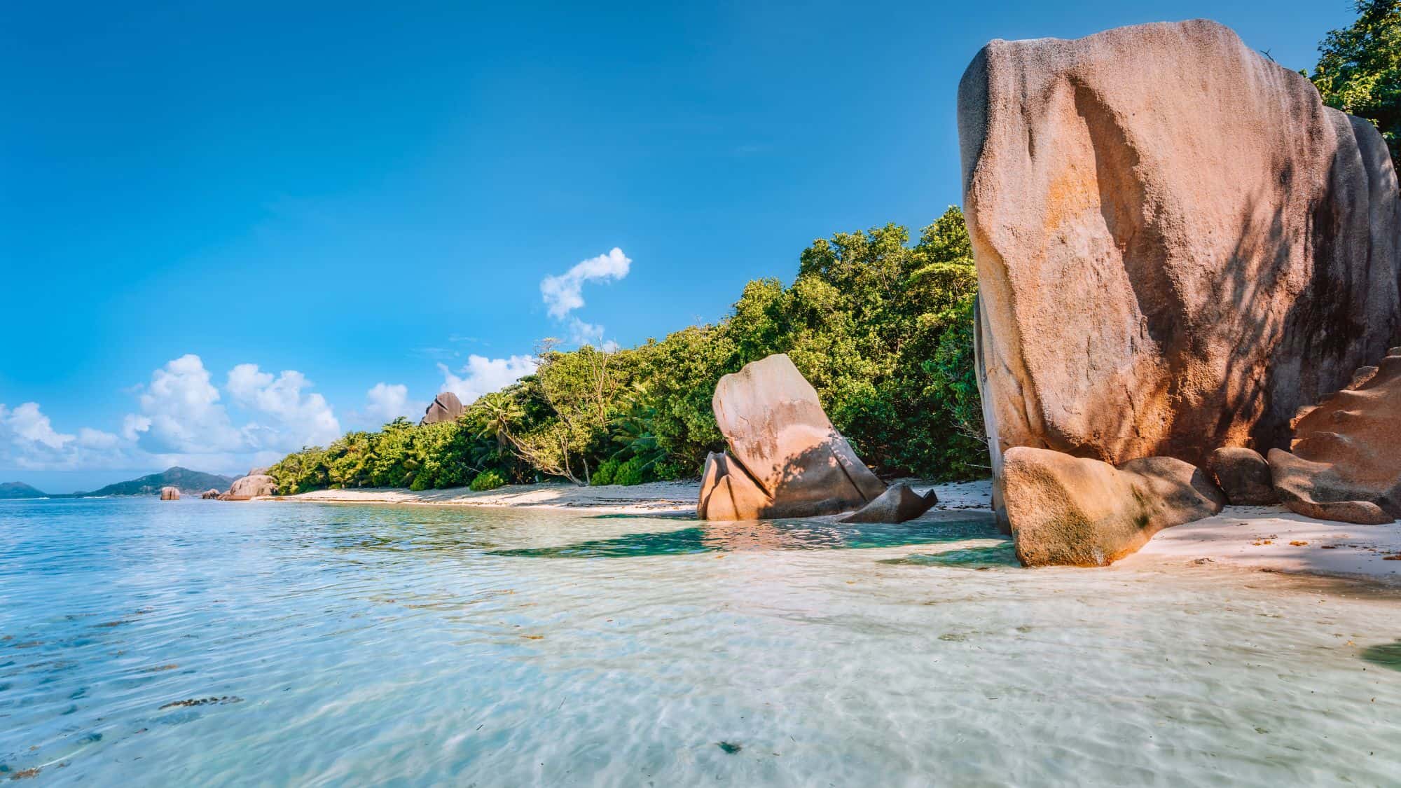 Towering granite boulders stand beside calm, clear shallows and a tree-lined shoreline on a sunny day.