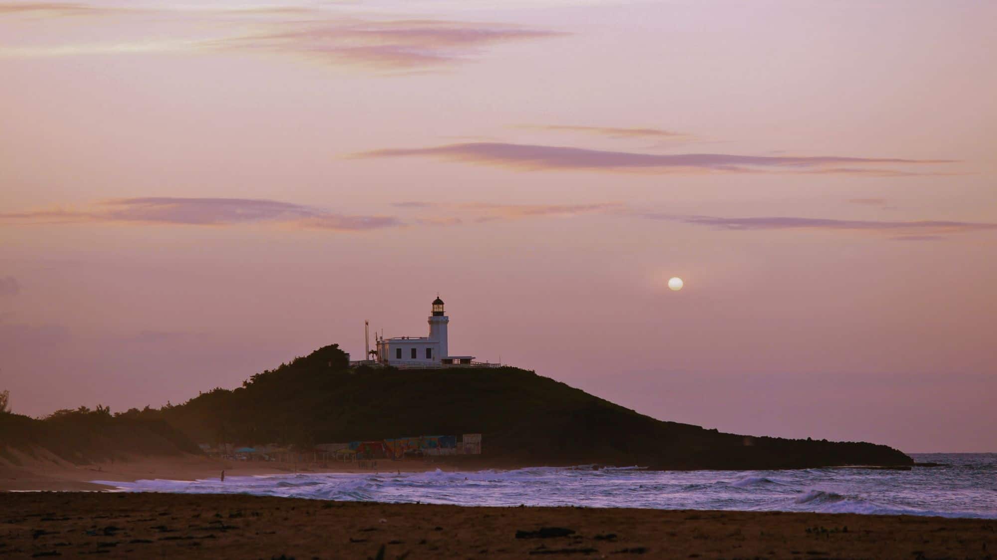 A white lighthouse sits atop a green hill along the coast of Aguadilla, Puerto Rico, silhouetted against a soft pink and purple sky with the sun low on the horizon and gentle waves rolling onto the beach.
