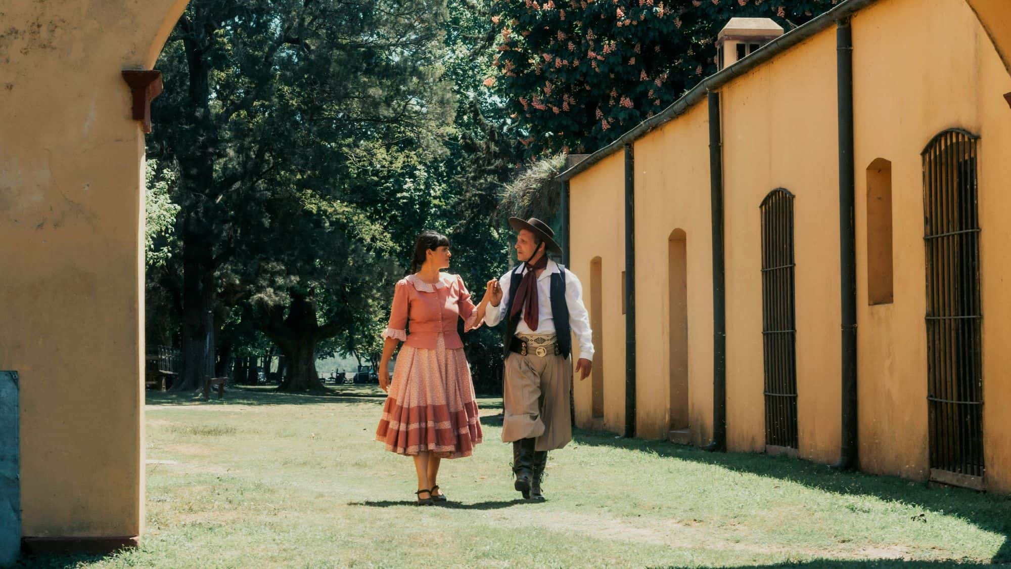A couple dressed in traditional Argentine attire walk hand in hand outside a rustic building, appearing mid-dance.