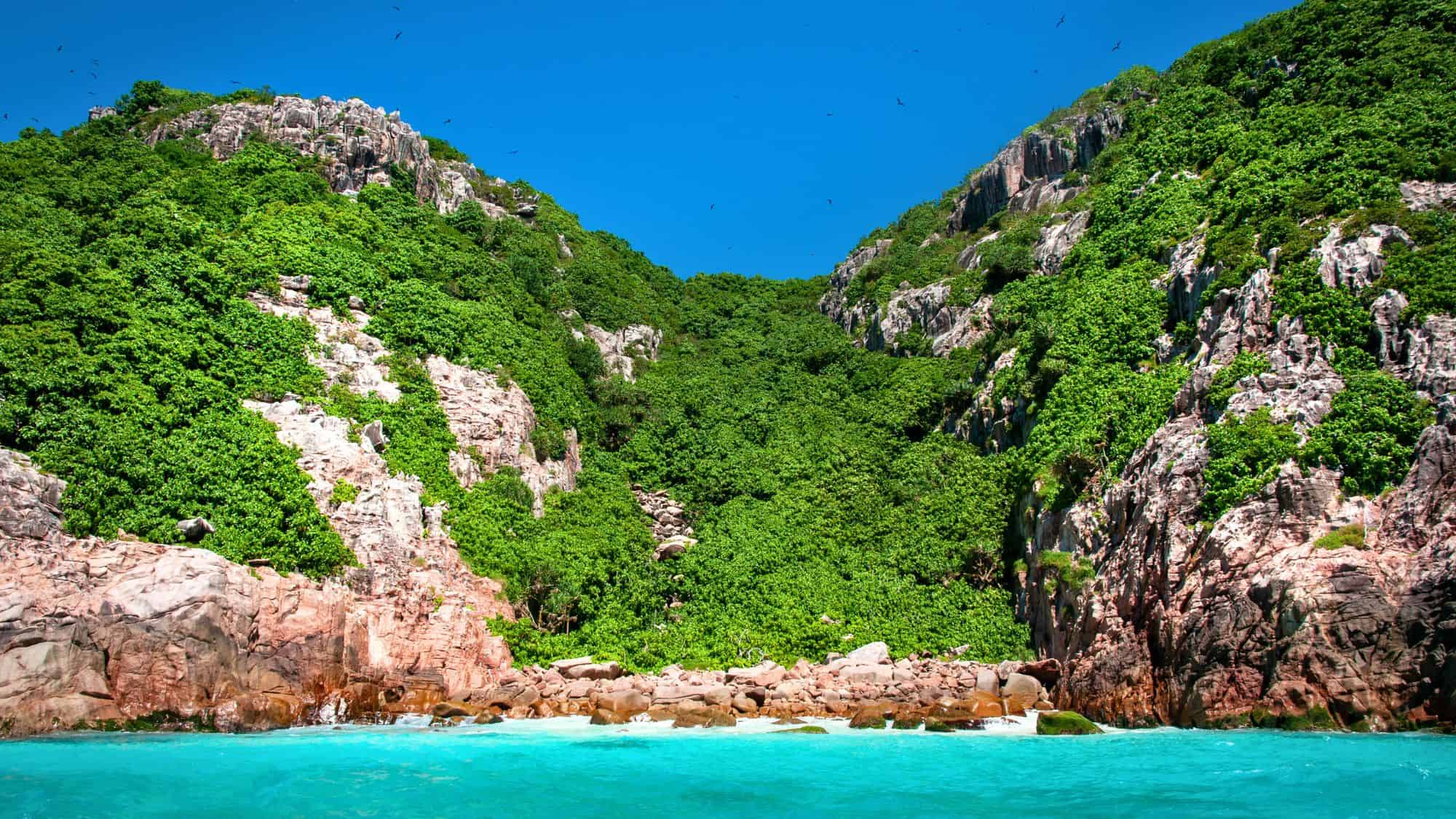 Steep rocky cliffs covered in dense green foliage rise above bright turquoise coastal waters under a clear blue sky.