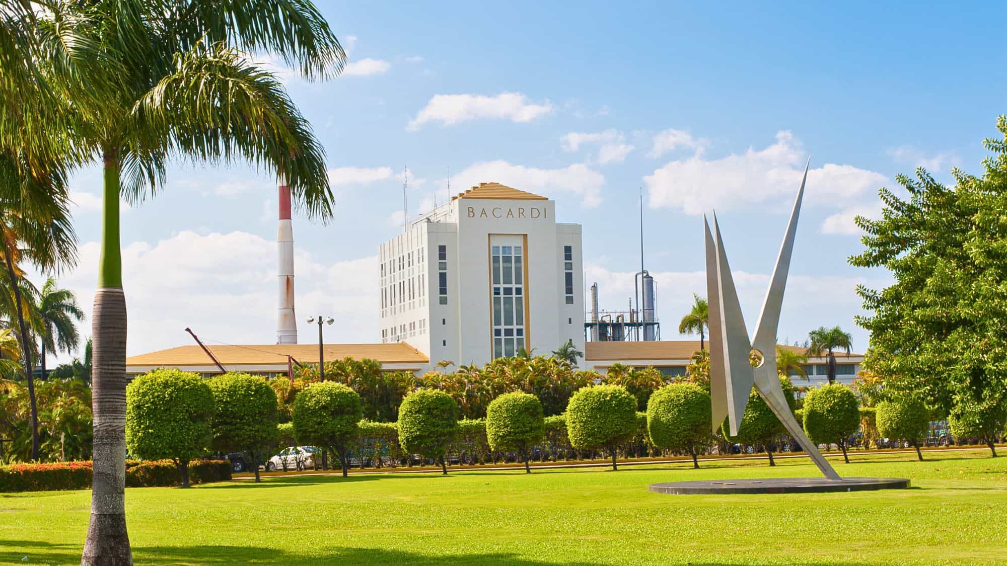 The Bacardi factory in Puerto Rico stands tall behind manicured lawns and palm trees, with a modern sculpture in the foreground under a clear blue sky.