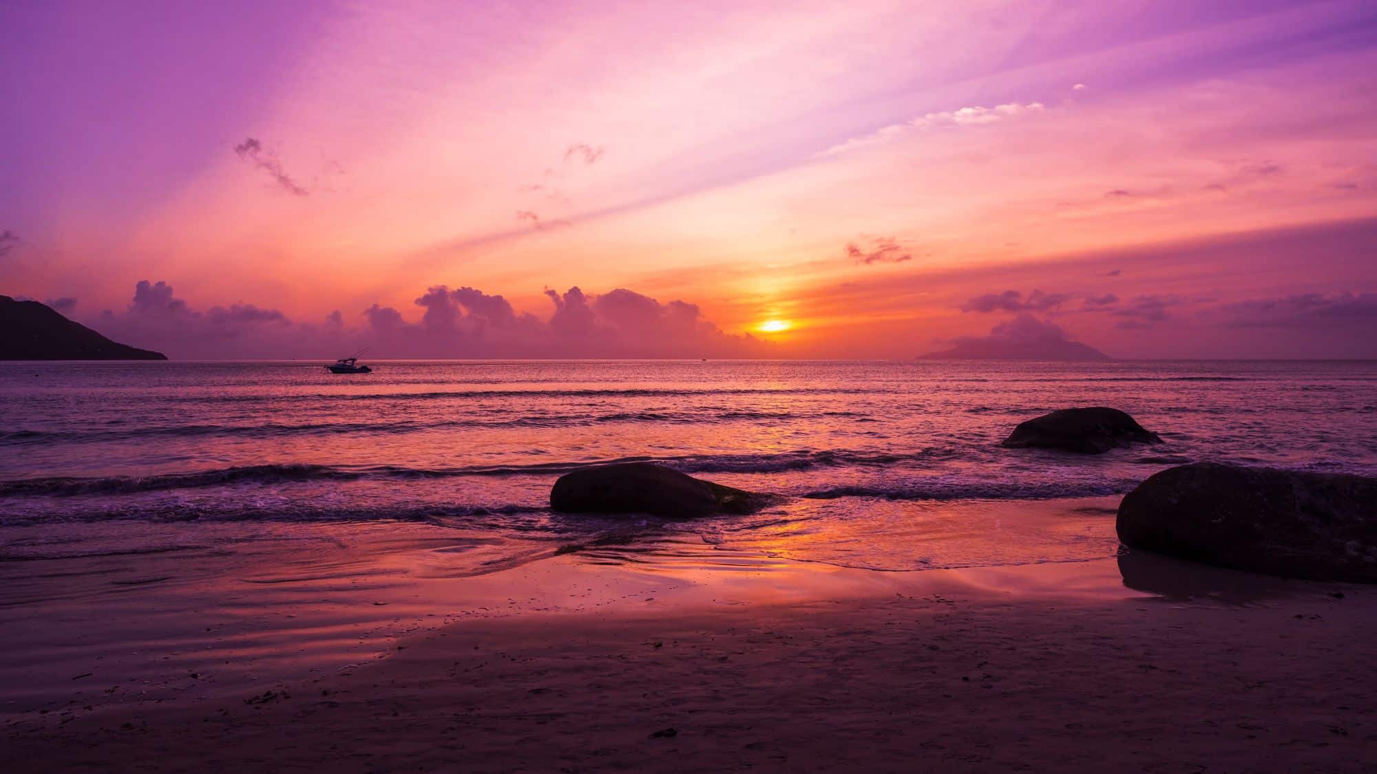 A fishing boat floats on calm seas at sunrise as the sky glows in pink, purple, and orange over a tranquil beach.