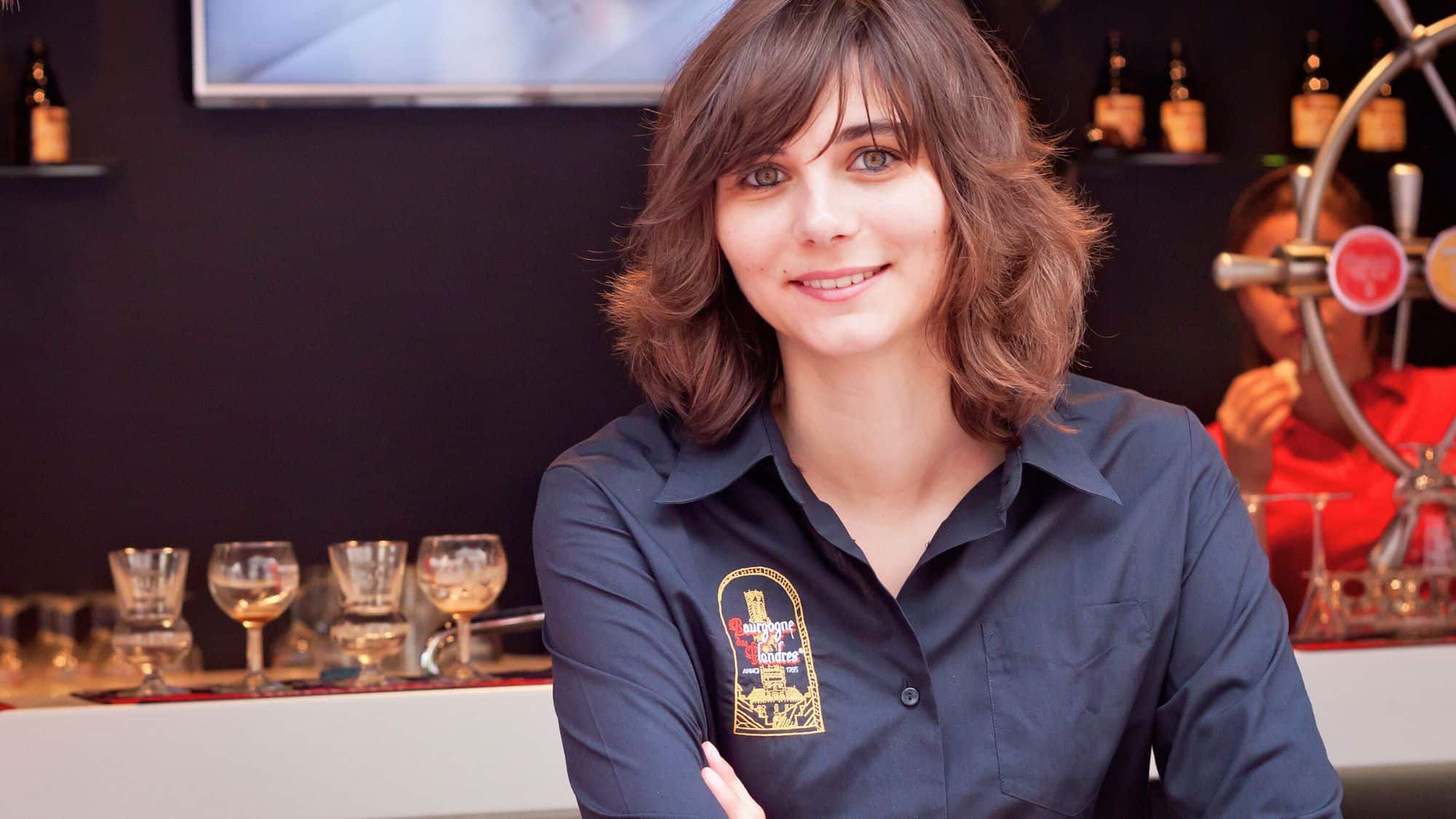 A smiling woman wearing a Bourgogne des Flandres shirt stands at a bar with beer taps and glasses in the background.