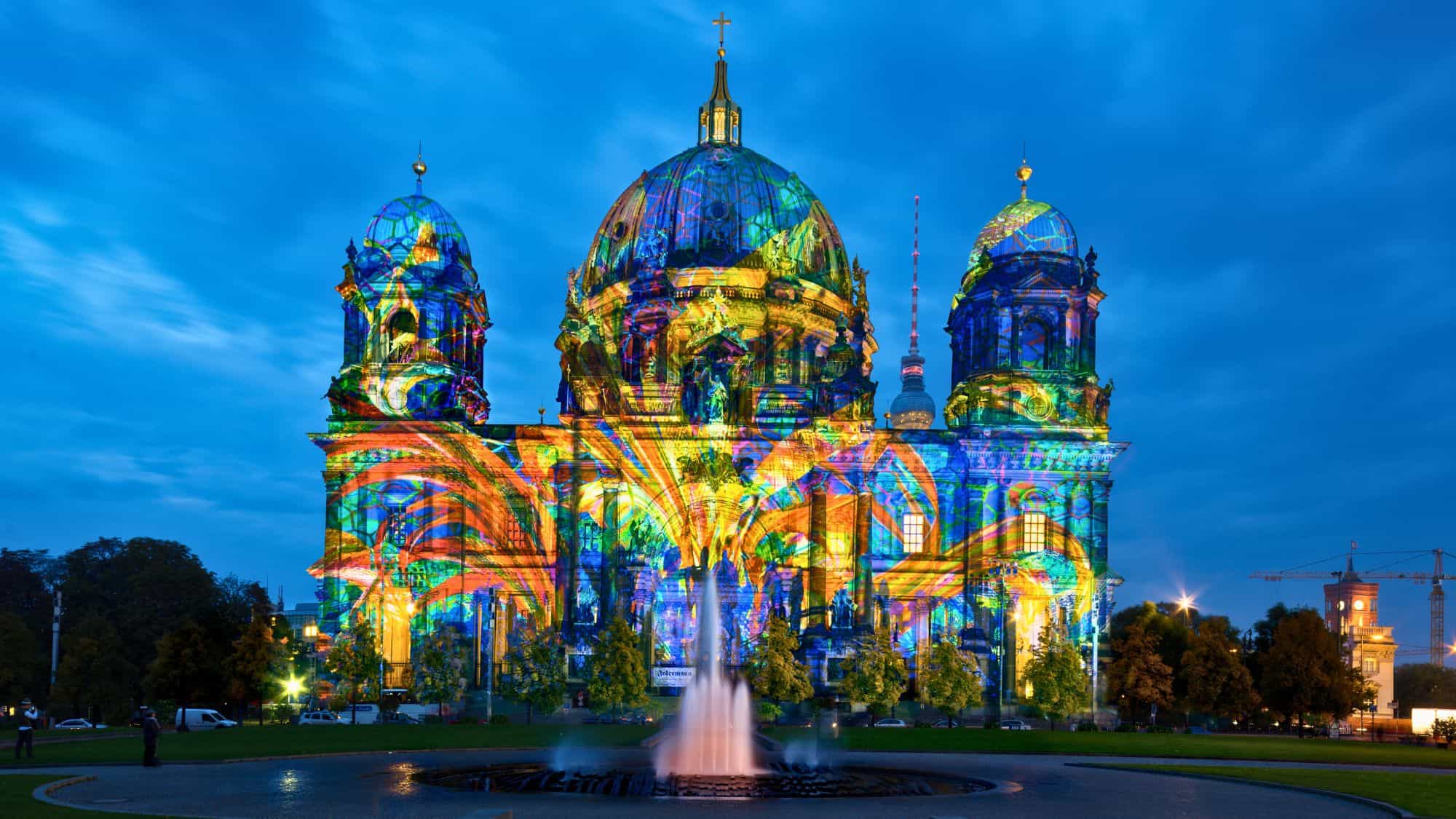 The Berlin Cathedral is covered in vivid, multicolored light projections that transform its domes and facade into a glowing canvas.
