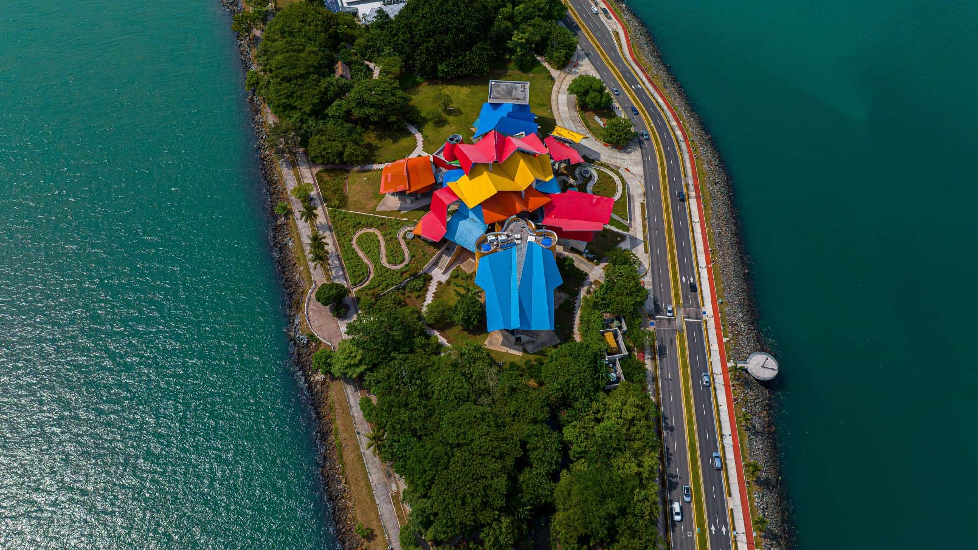 The Biomuseo in Panama City features a striking design of overlapping, colorful angular roofs on a narrow strip of land surrounded by bright blue water and a coastal highway.
