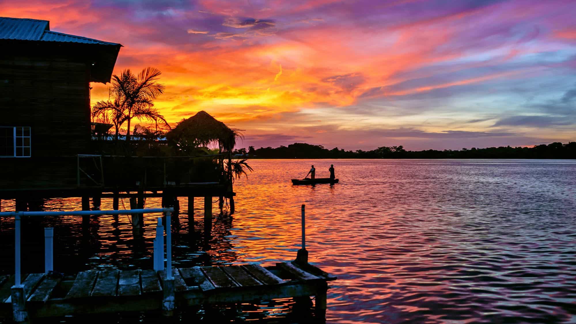 Two figures paddle a small boat across the still waters of Bocas del Toro, Panama, under a dramatic sunset sky filled with oranges, purples, and pinks.