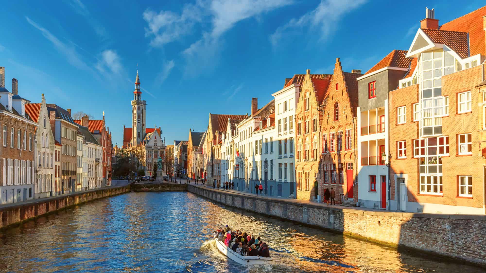 A boat filled with passengers glides through a canal lined with colorful historic buildings in Bruges, Belgium.