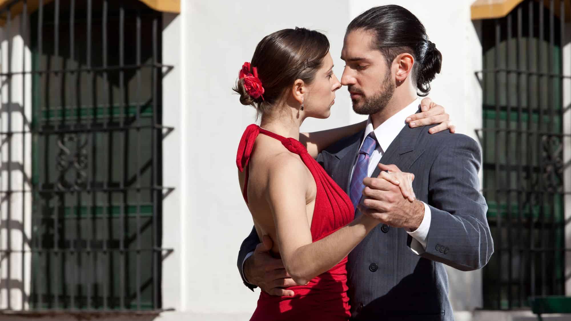A couple locked in a passionate tango pose, the woman in a red dress and the man in a suit, gazing intently at each other.