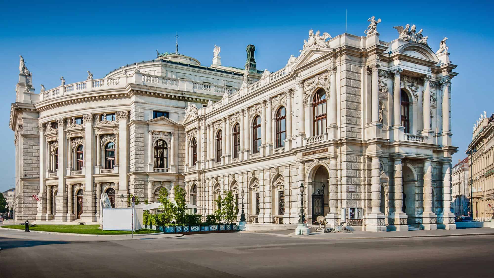 A grand neoclassical theater with ornate statues and arched windows stands under a bright blue sky in Vienna.