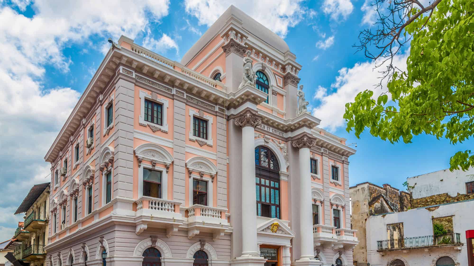A neoclassical pink and white government building with grand columns and statues stands out in Panama City's historic Casco Viejo under a partly cloudy sky.
