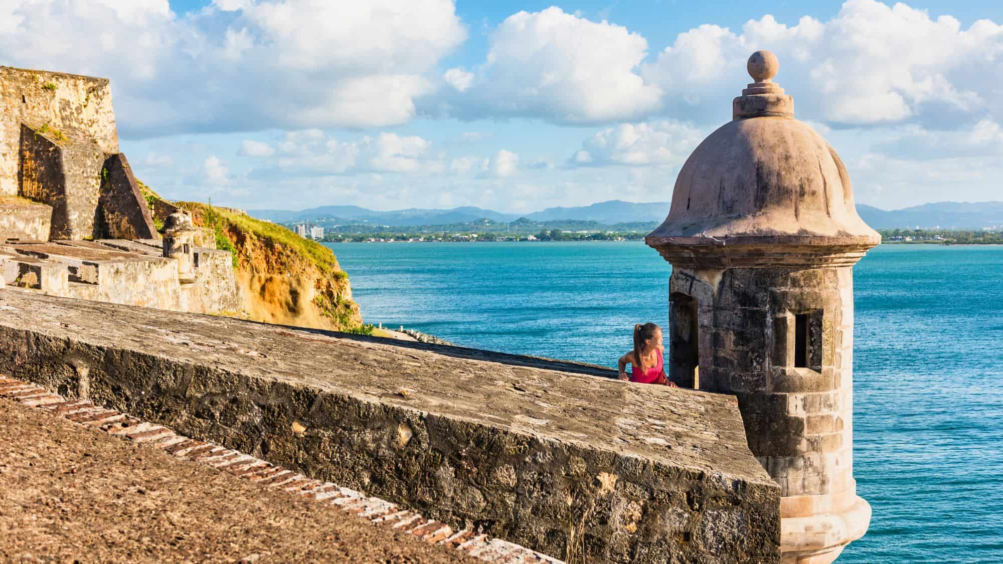 A woman explores the historic stone walls and garita (sentry box) of Castillo San Felipe del Morro, overlooking the bright blue waters of San Juan Bay.