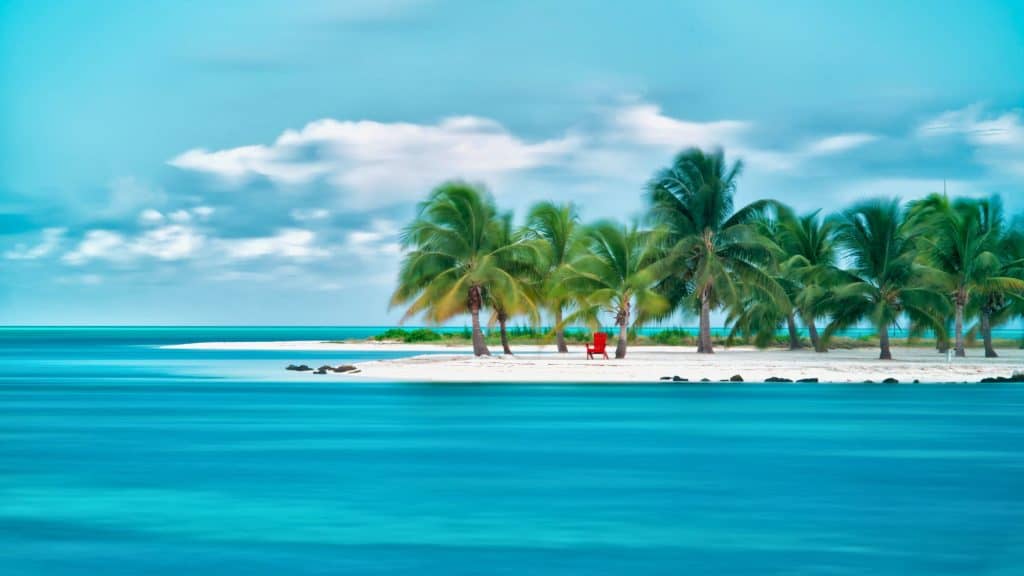 A tiny tropical island lined with palm trees features a single bright red chair facing the turquoise sea.