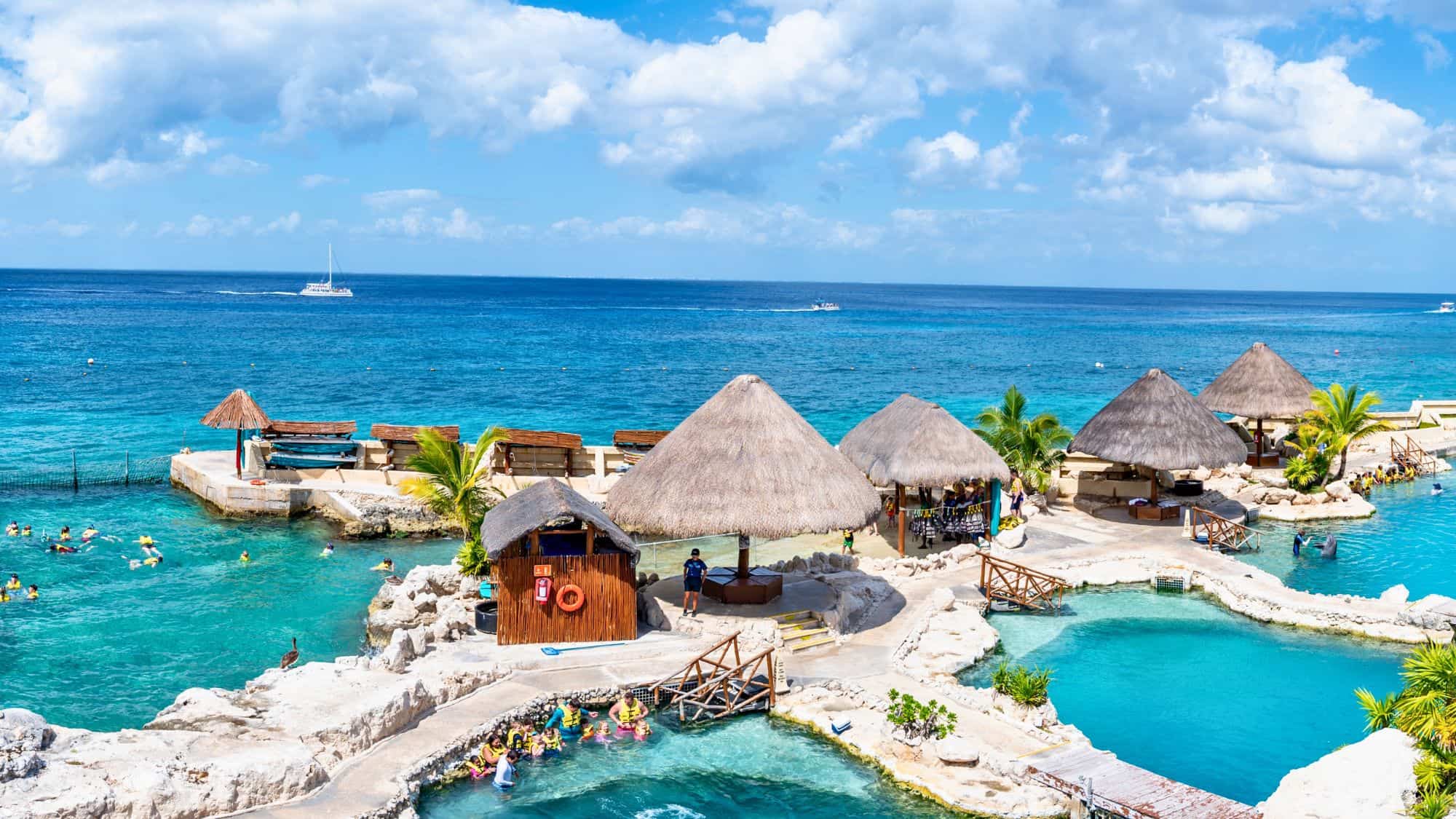 Thatched-roof cabanas and turquoise pools overlook the bright blue Caribbean Sea, with snorkelers swimming nearby.