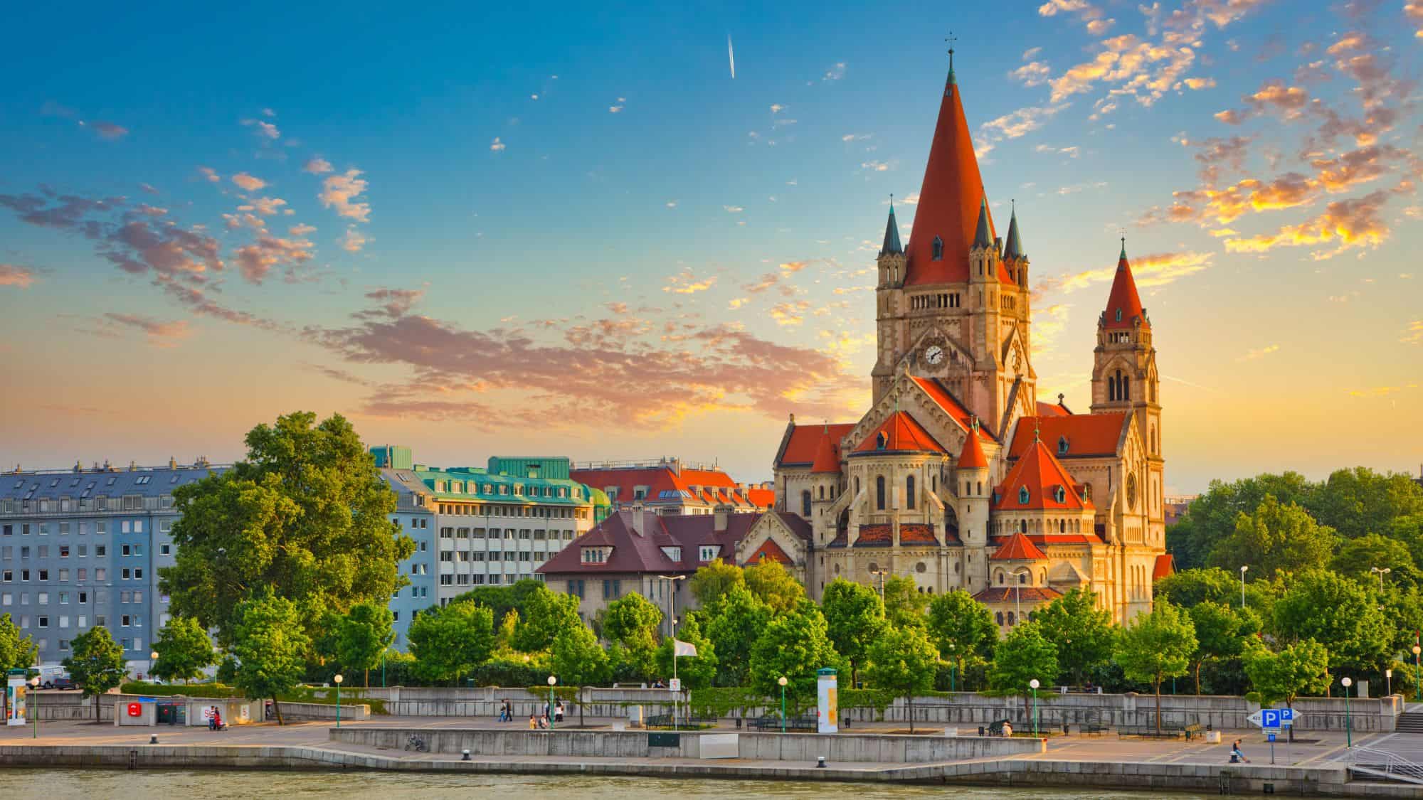 A striking red-roofed church with tall pointed towers rises above the Danube River against a golden sunset sky.
