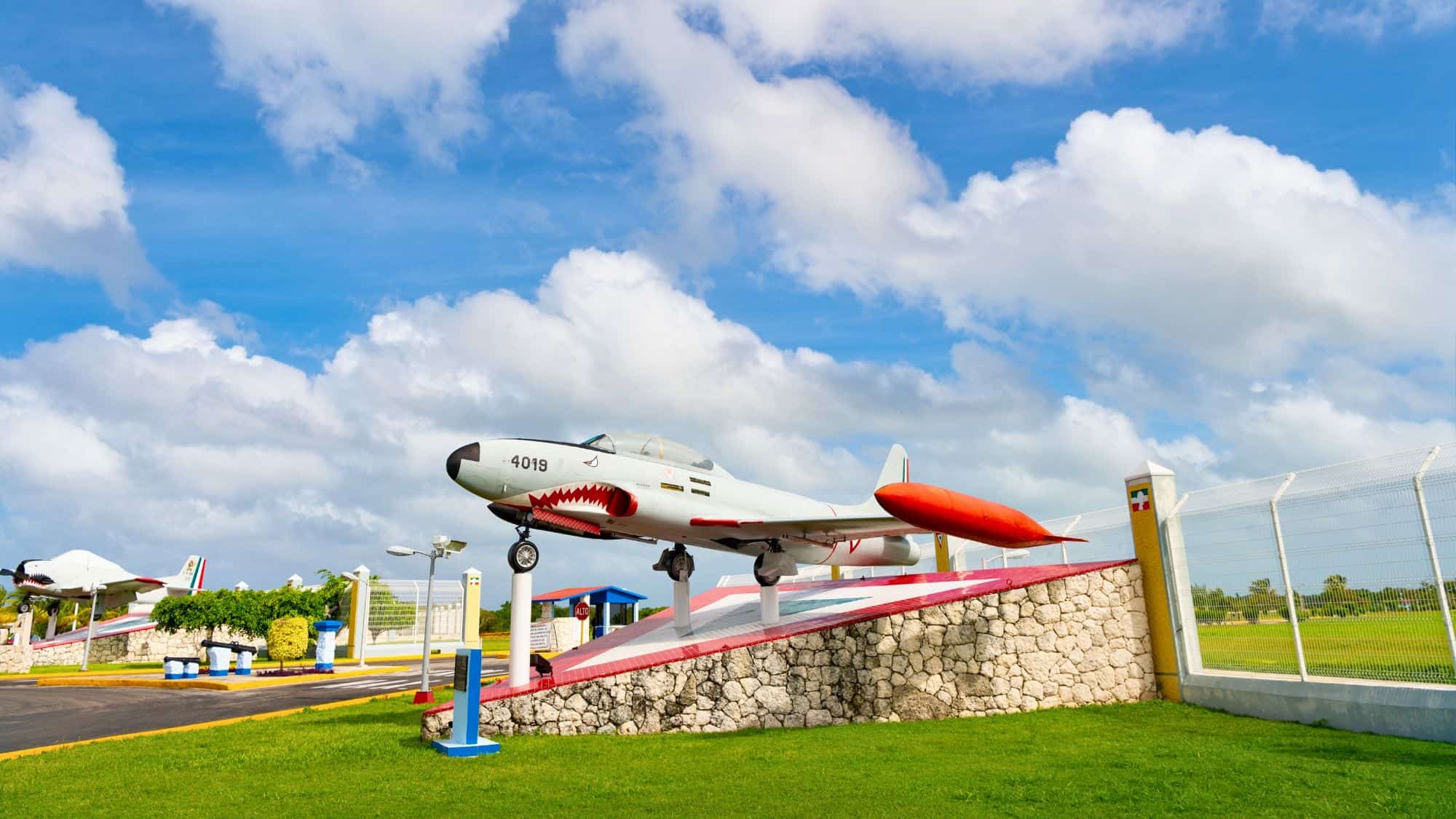 A vintage fighter jet painted with a shark mouth design is mounted on a stone platform at an outdoor exhibit.