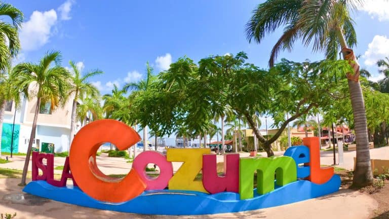 A large, colorful “Isla Cozumel” sign sits among palm trees in a sunny plaza with a bright blue sky overhead.