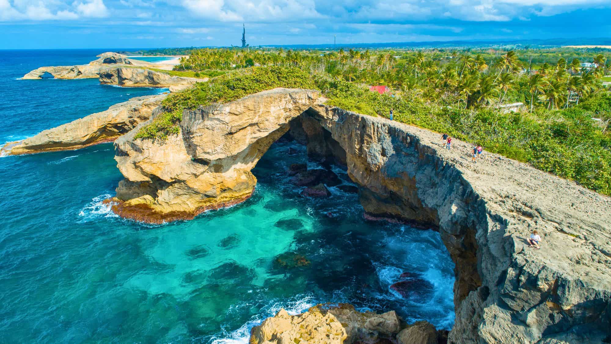 Dramatic limestone cliffs and a natural arch rise over vibrant turquoise waters at Cueva del Indio, where visitors walk along the edge of the rocky coastal landscape.