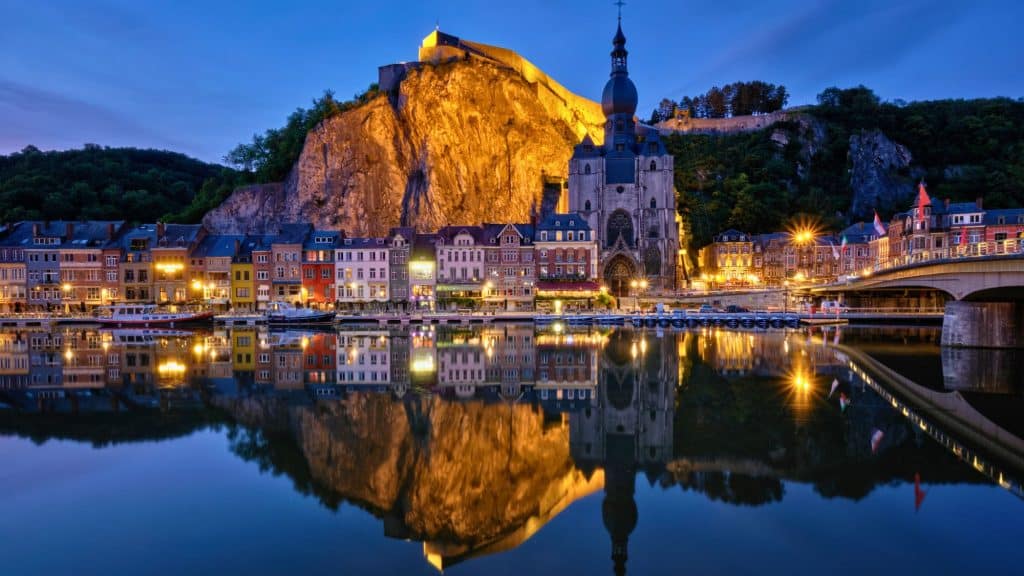 The illuminated Collegiate Church of Notre Dame and colorful riverside buildings reflect in the calm waters beneath a rocky cliff in Dinant, Belgium.