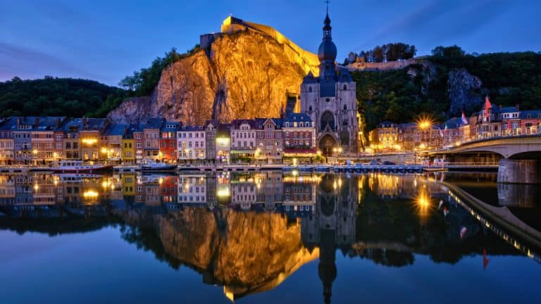 The illuminated Collegiate Church of Notre Dame and colorful riverside buildings reflect in the calm waters beneath a rocky cliff in Dinant, Belgium.
