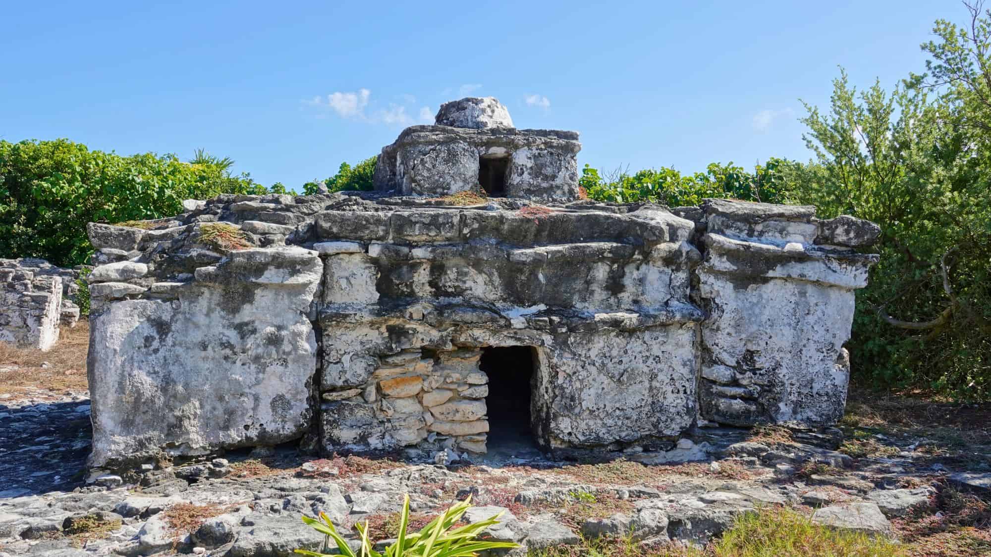 A weathered stone Mayan structure with small entryways stands surrounded by lush greenery under a clear sky.