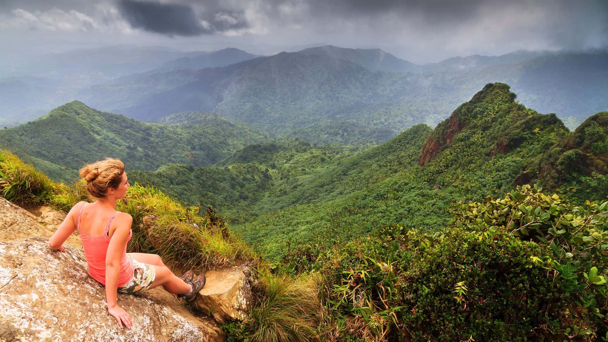 A hiker sits on a rock, taking in the lush green mountains of El Yunque National Forest under a cloudy sky, with mist drifting over the peaks.