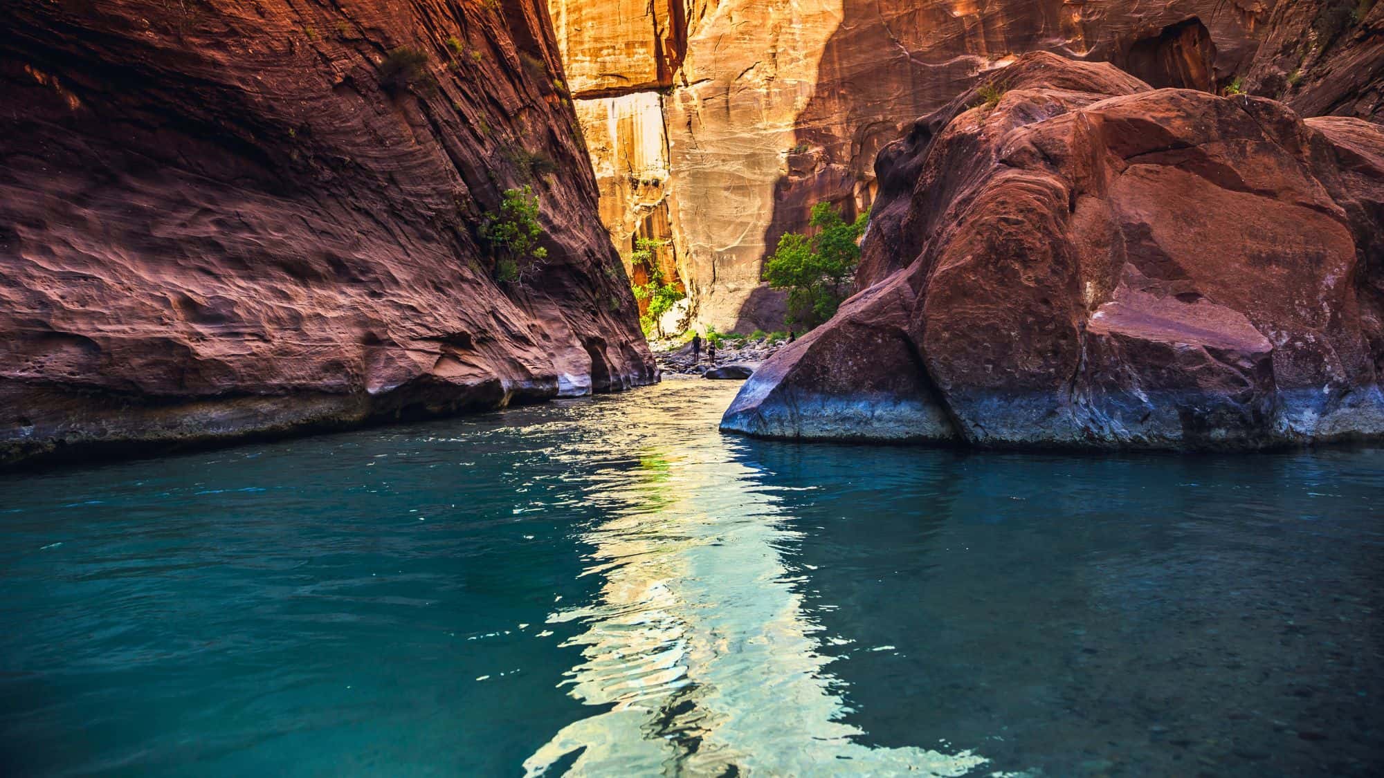 Sunlight reflects off turquoise water flowing between towering red rock canyon walls, with patches of greenery along the edges.
