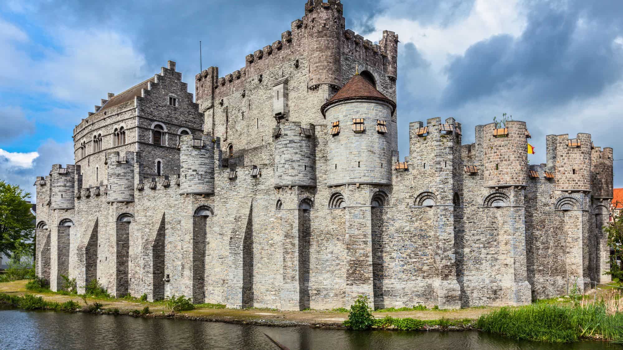The medieval stone fortress of Gravensteen stands tall with turrets and battlements, surrounded by a moat in Ghent, Belgium.