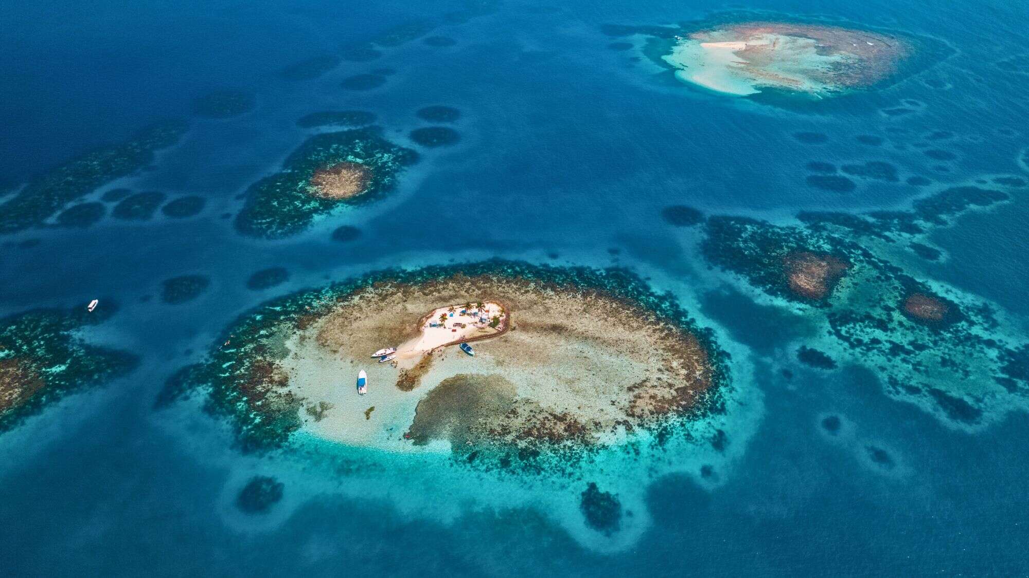 A tiny sandy island surrounded by turquoise waters and coral reefs, with a few boats anchored nearby.
