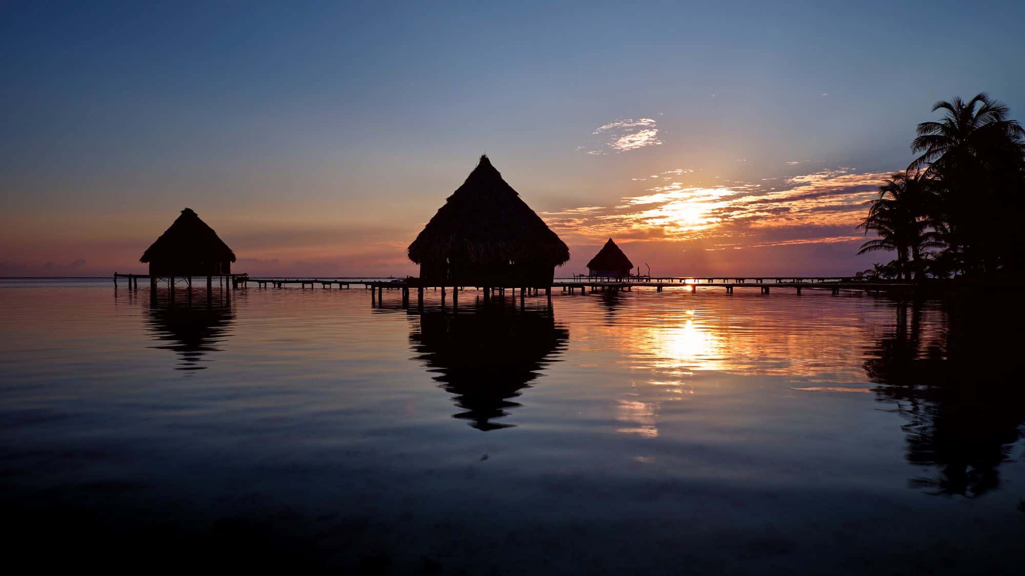 Silhouetted thatched-roof cabanas on stilts over calm water, reflecting a vivid tropical sunset.