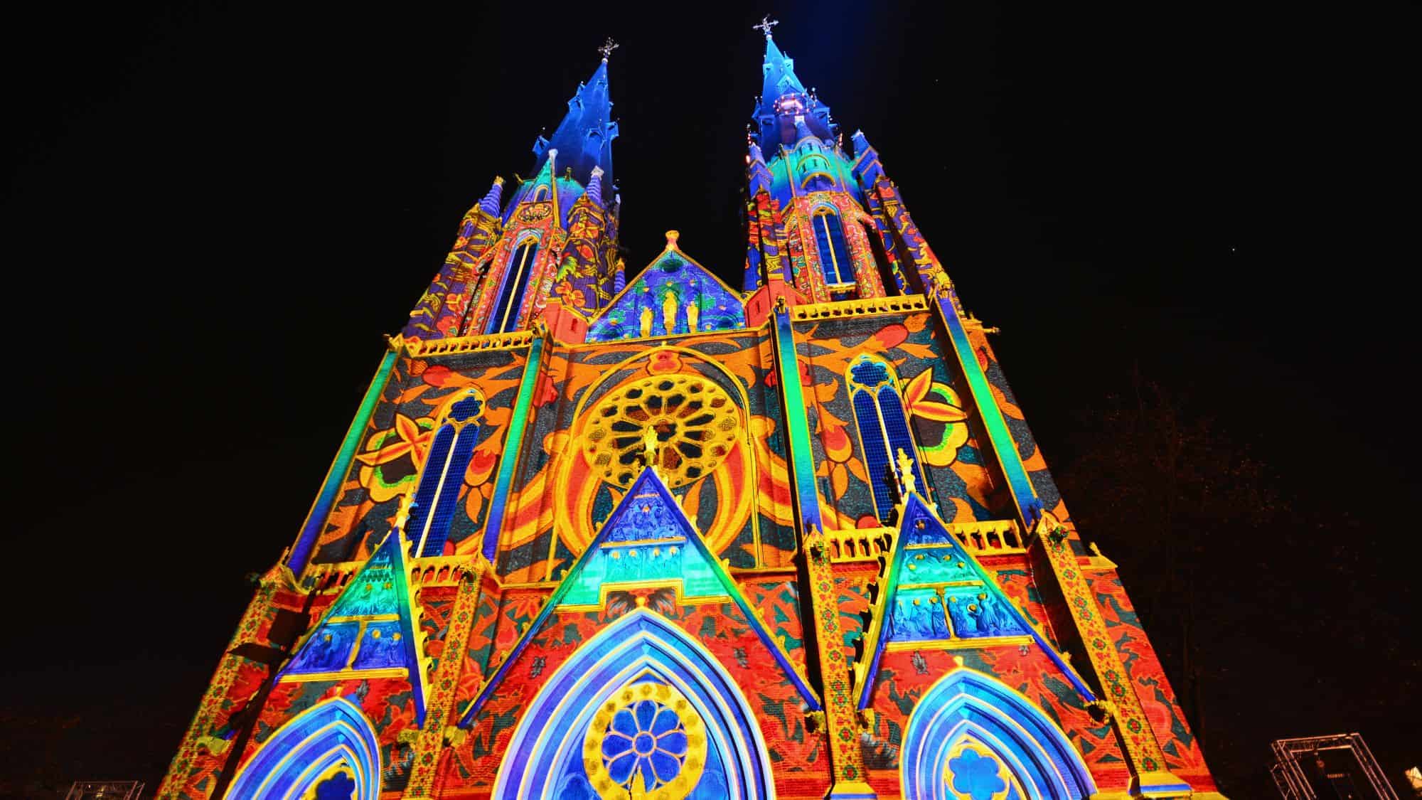 A Gothic cathedral is bathed in vivid, multicolored light projections that highlight its towers and stained-glass windows.