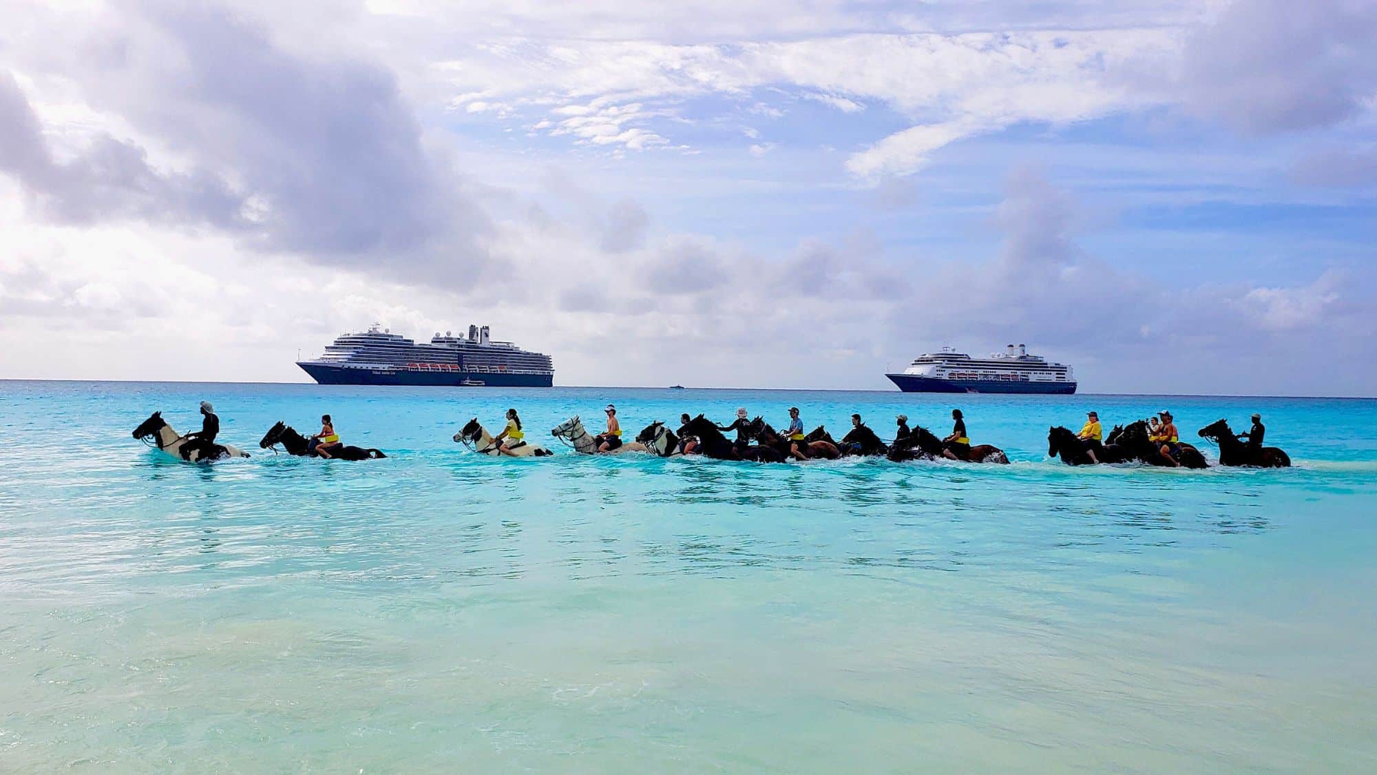 A group of riders guiding horses through shallow turquoise waters, with cruise ships anchored in the distance.