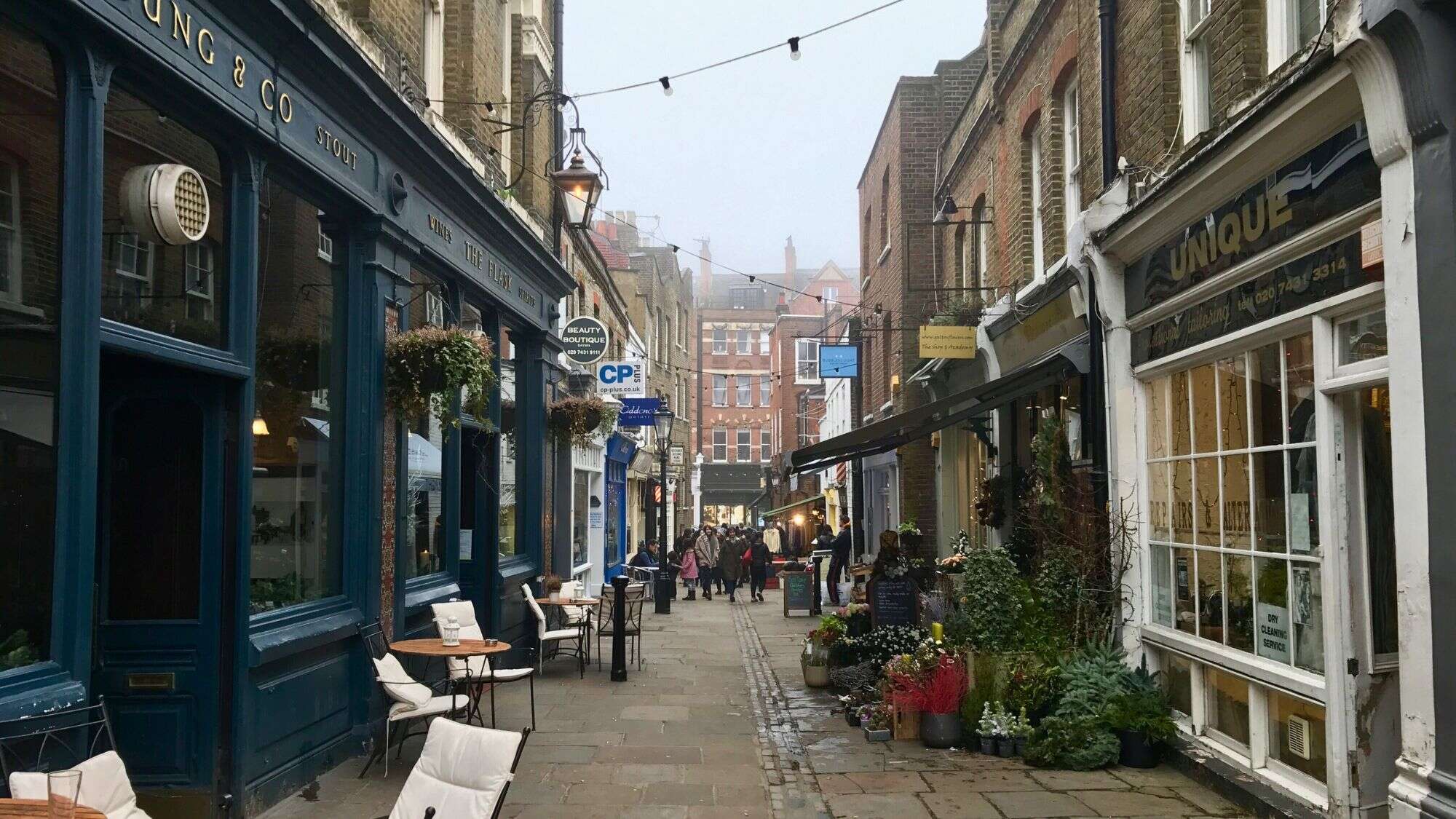 A narrow cobblestone street lined with cozy cafés, boutique shops, and hanging plants, with people strolling under string lights.