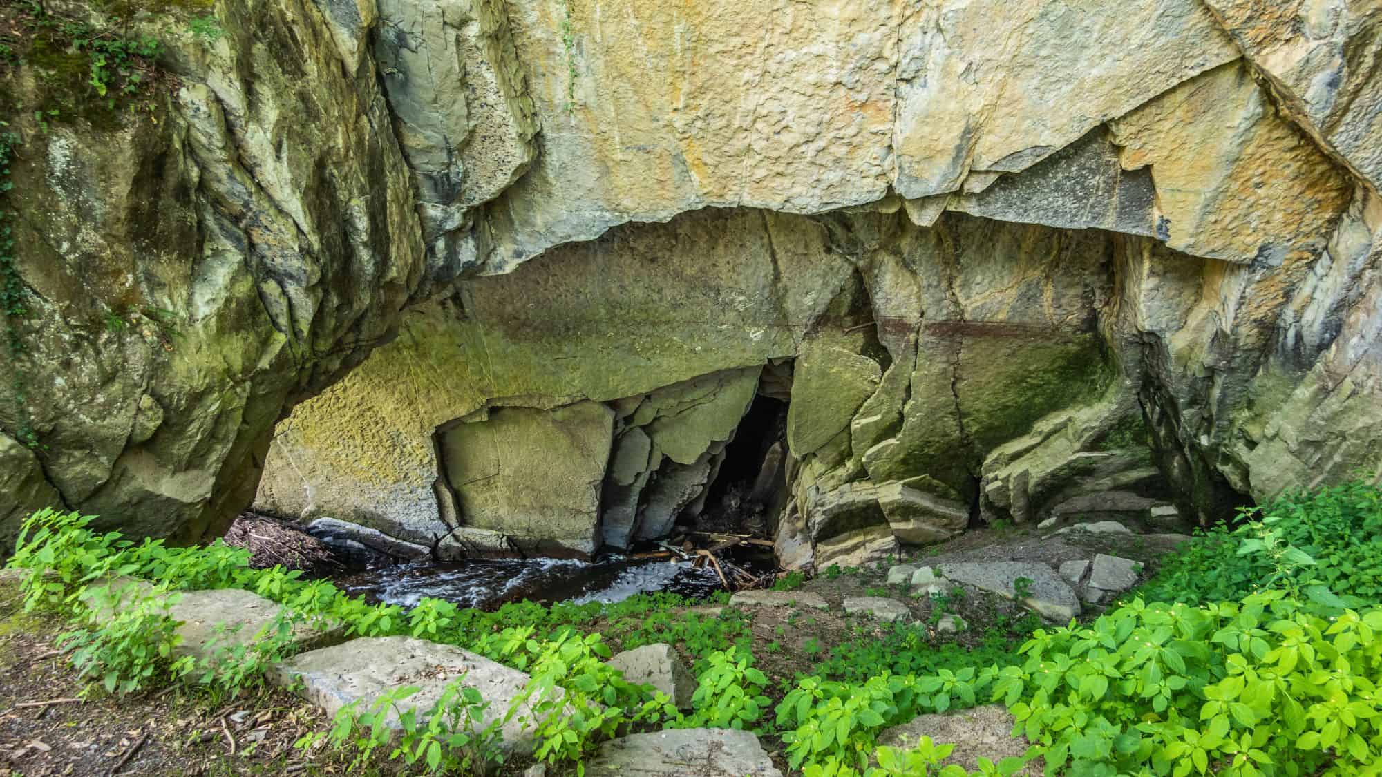 The rocky entrance to the Caves of Han features jagged stone formations above a small flowing stream.